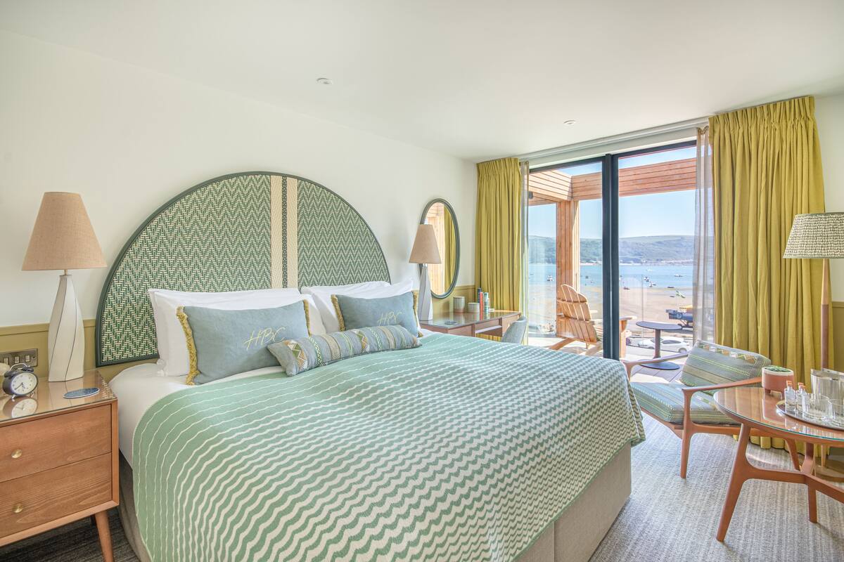 A hotel room with a large bed, a desk with a chair, a table with a chair, and a view of the sea.