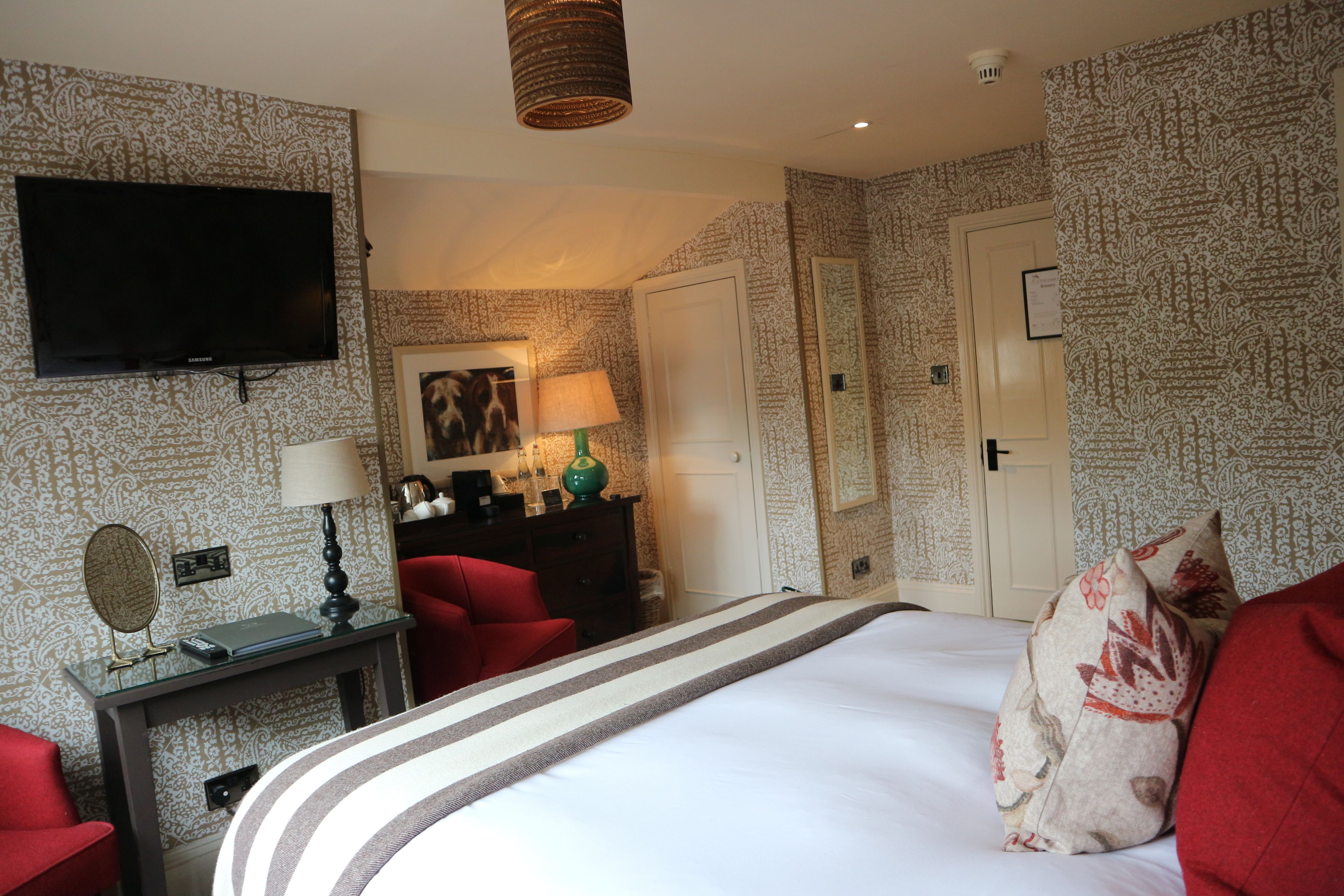 A hotel room with a large bed, a television mounted on the wall, a desk with a lamp, and a red chair.