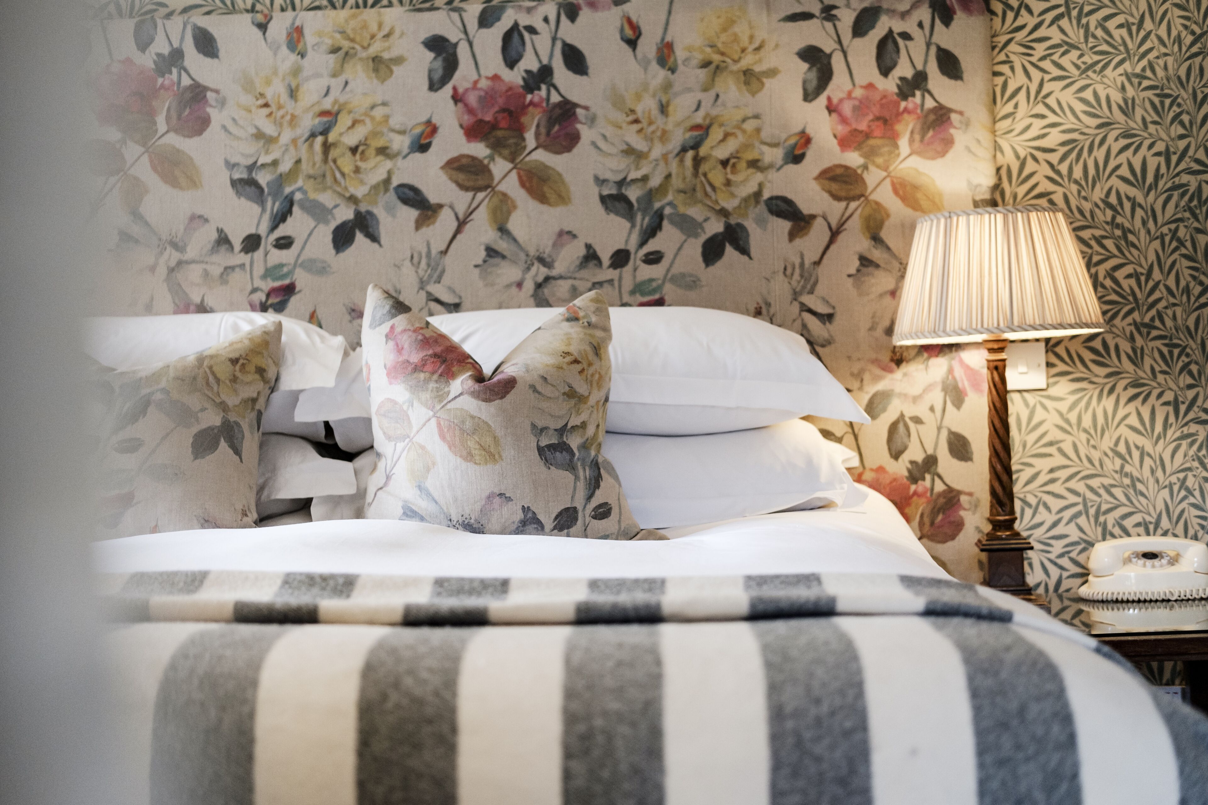 A bed with floral-patterned pillows, a striped blanket, and a bedside table with a lamp and a rotary phone.