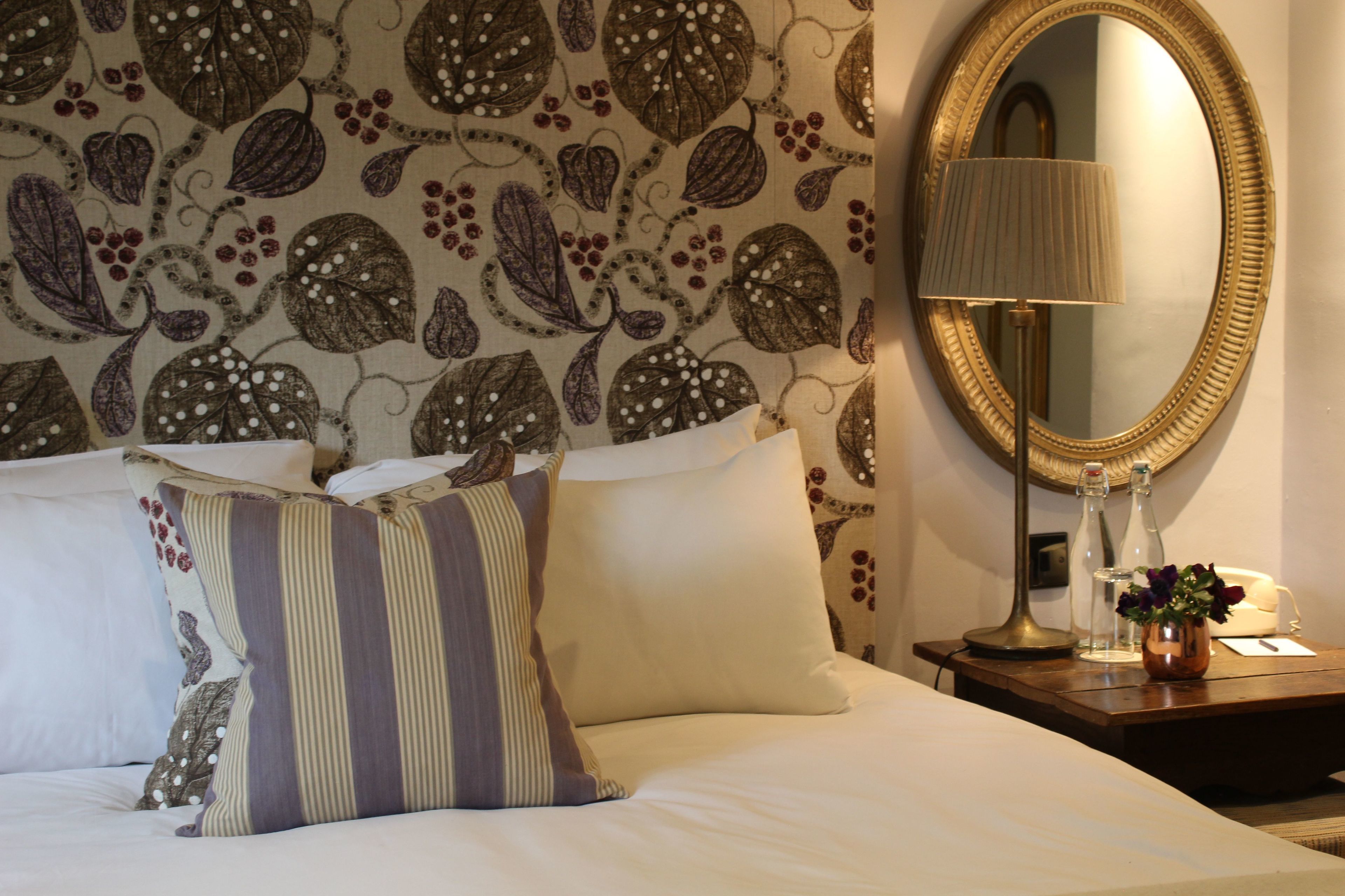 A hotel room with a bed, pillows, a nightstand with a lamp, a vase with flowers, and a mirror on the wall.