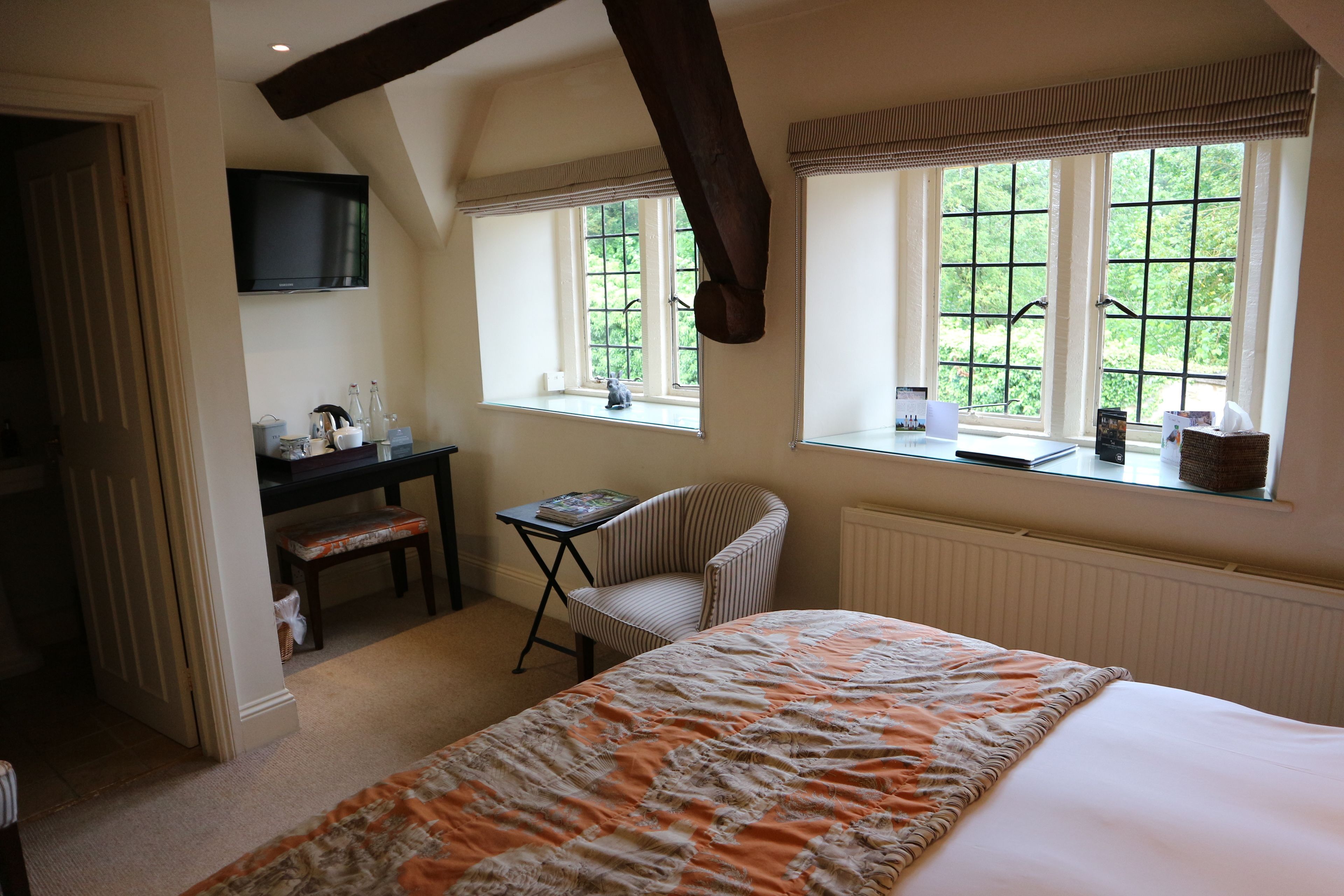 A bedroom with a bed, a desk, a chair, and two windows with a view of greenery.