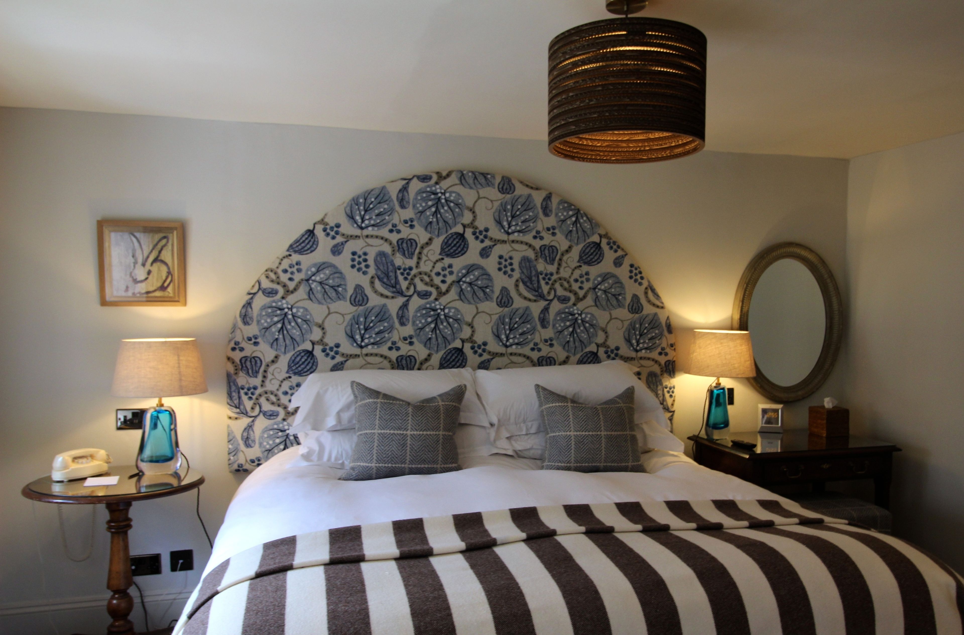 A hotel room with a large floral-patterned headboard, two bedside tables with lamps, a bed with striped bedding, and a mirror on the wall.