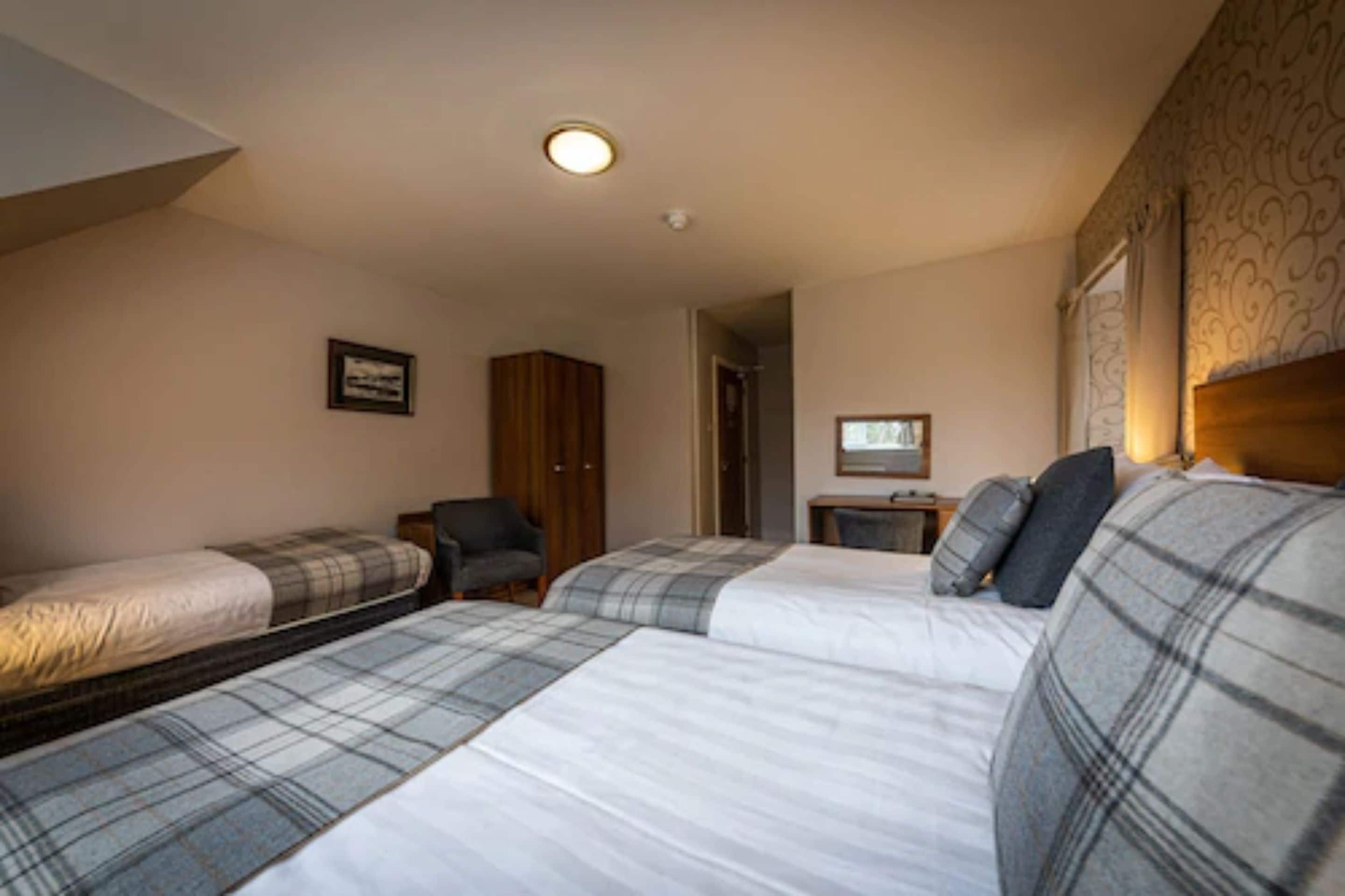 A room with three beds, a wooden wardrobe, a chair, and a small table with a lamp.