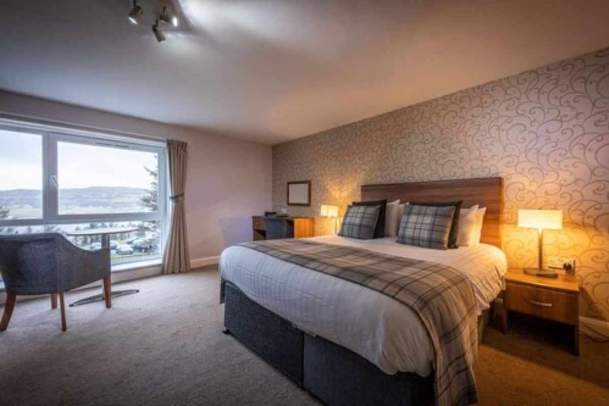 Quarto de hotel com uma cama grande, duas mesas de cabeceira, uma cadeira e vista para o exterior.