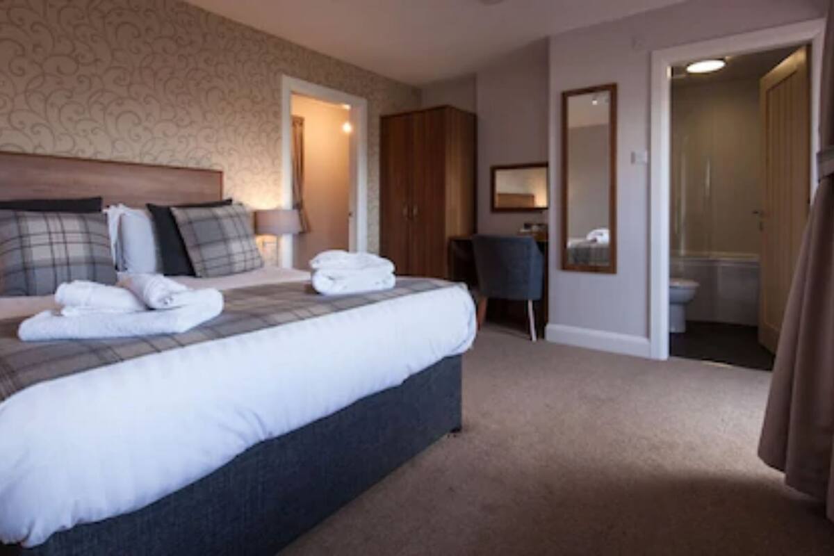 Quarto de hotel com uma cama grande, uma escrivaninha, um guarda-roupa e um banheiro visível através de uma porta aberta.