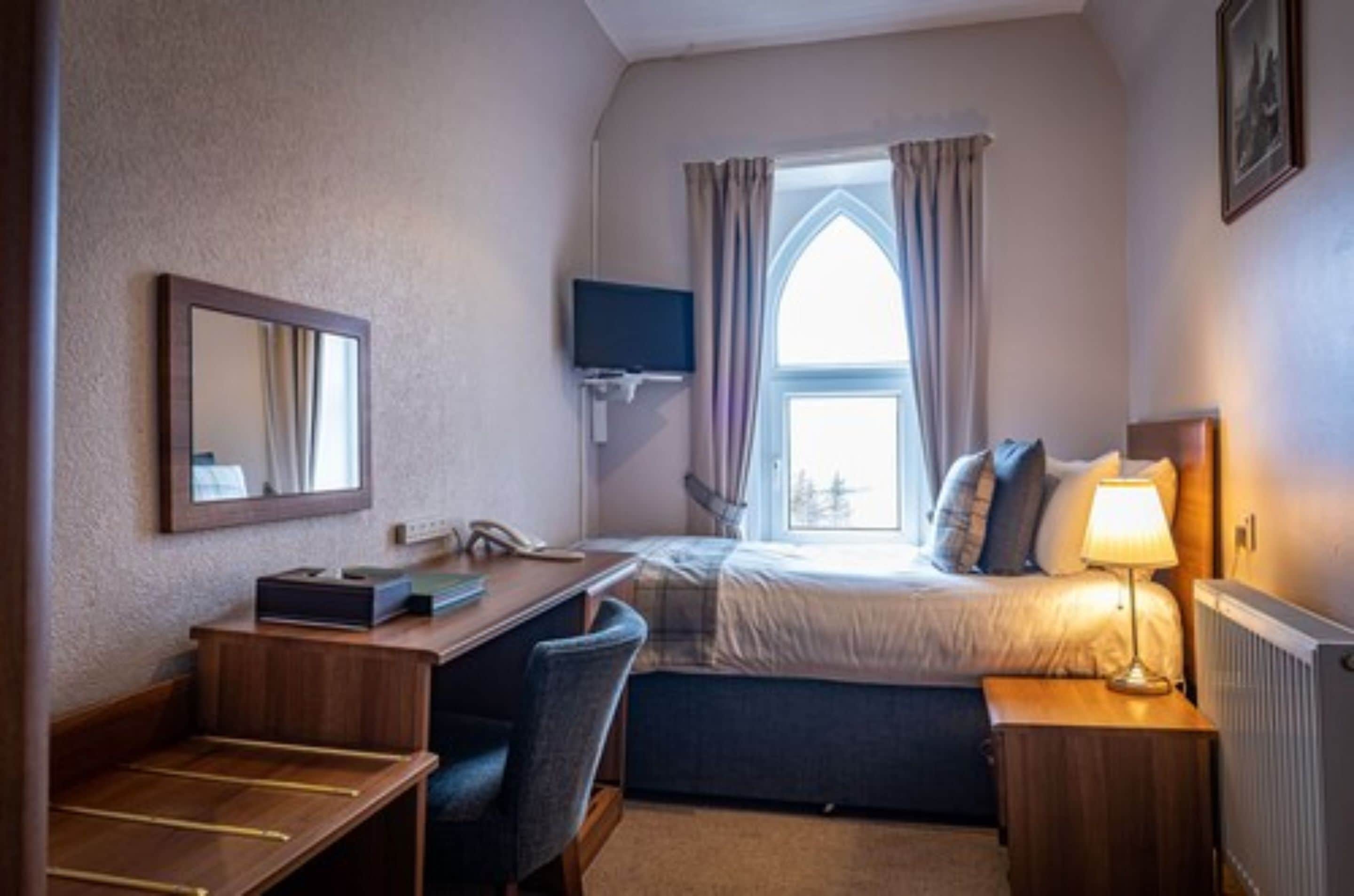A small hotel room with a single bed, a desk with a chair, a television mounted on the wall, and a window with curtains.