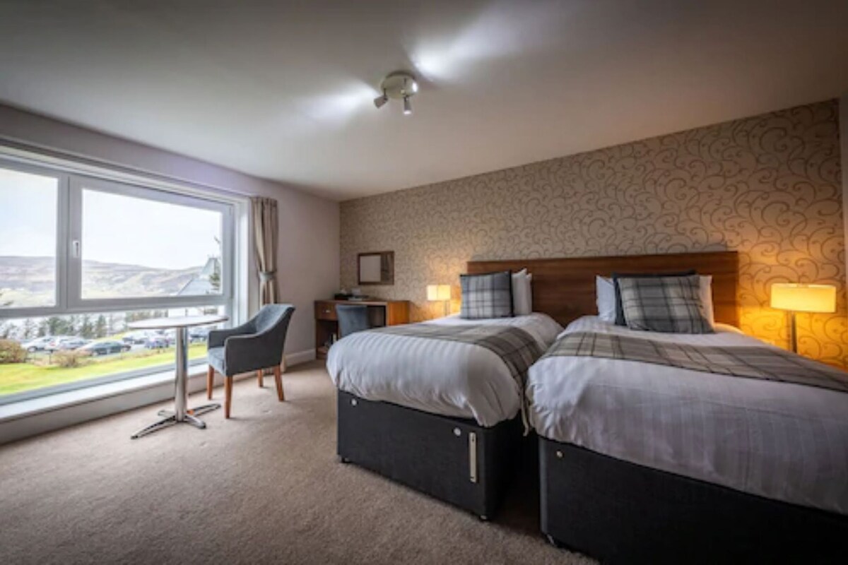 A hotel room with two beds, a desk, a chair, and a large window offering a view.
