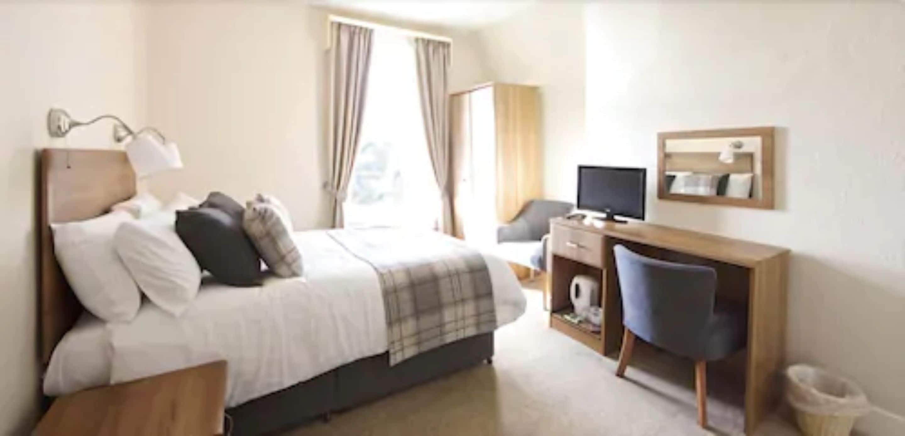 A hotel room with a bed, a desk, a chair, a TV, and a window with curtains.