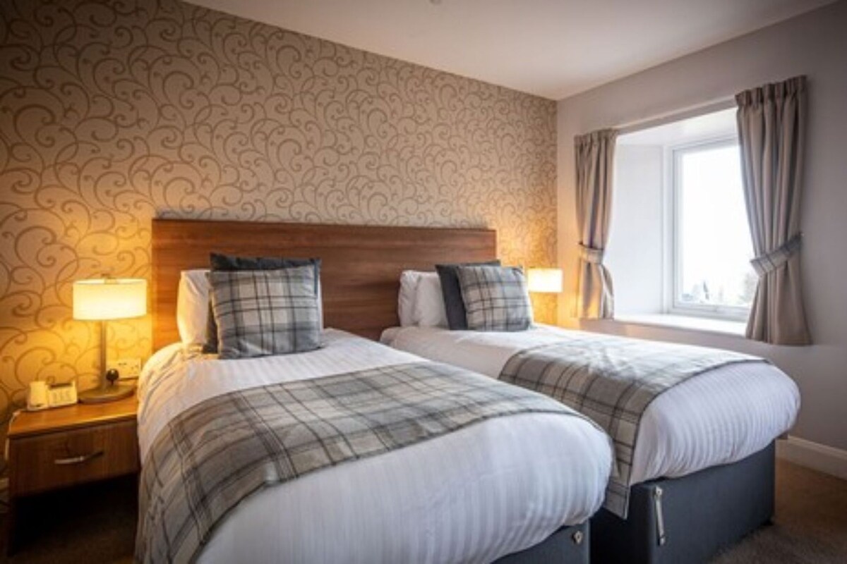 A hotel room with two beds, a wooden headboard, a nightstand with a lamp, and a window with curtains.