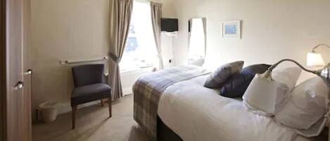 A hotel room with a bed, a chair, a bedside table with a lamp, a window with curtains, and a wall-mounted TV.