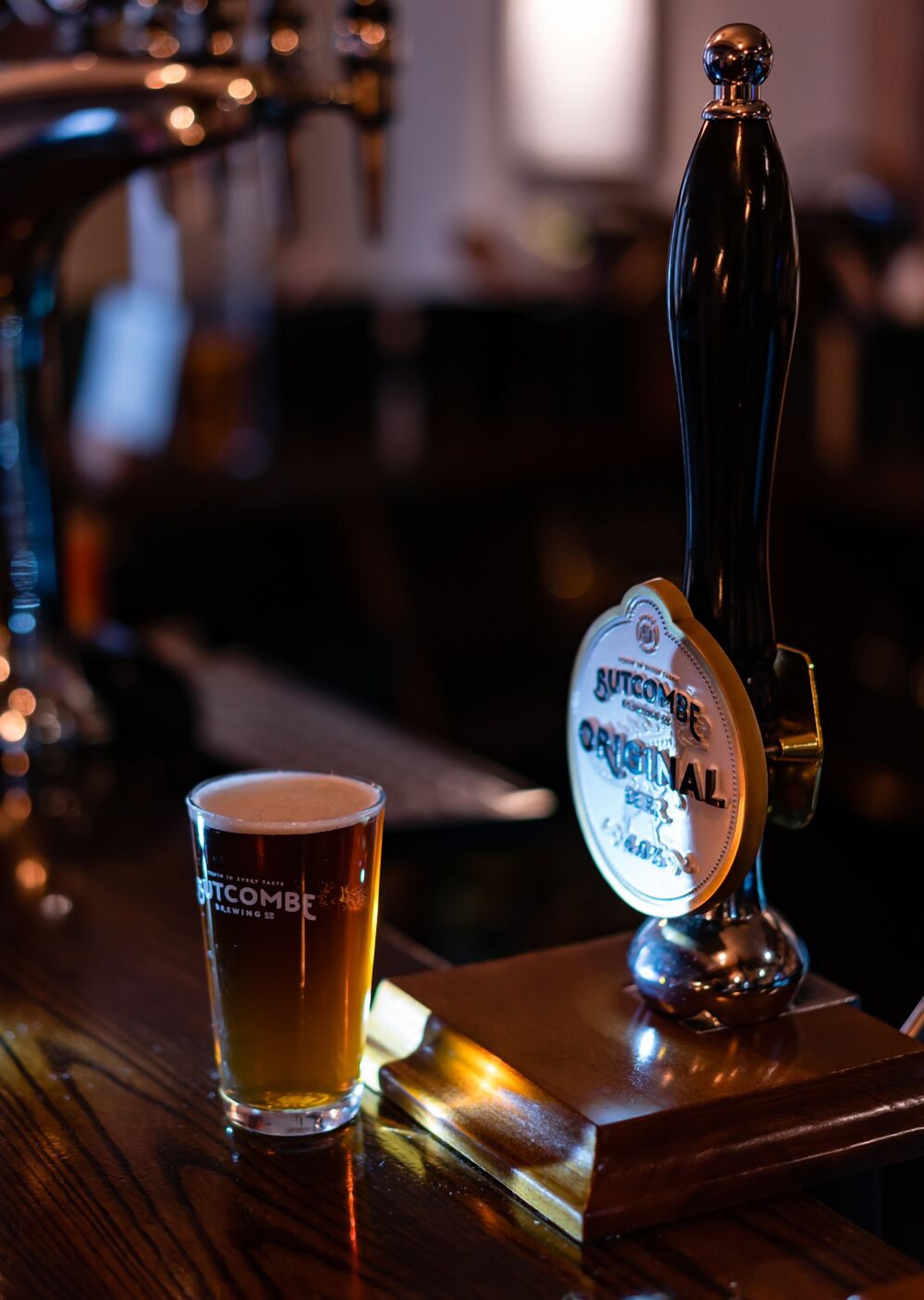 Un grifo con la etiqueta 'Butcombe Original' y un vaso de cerveza sobre una superficie de madera.