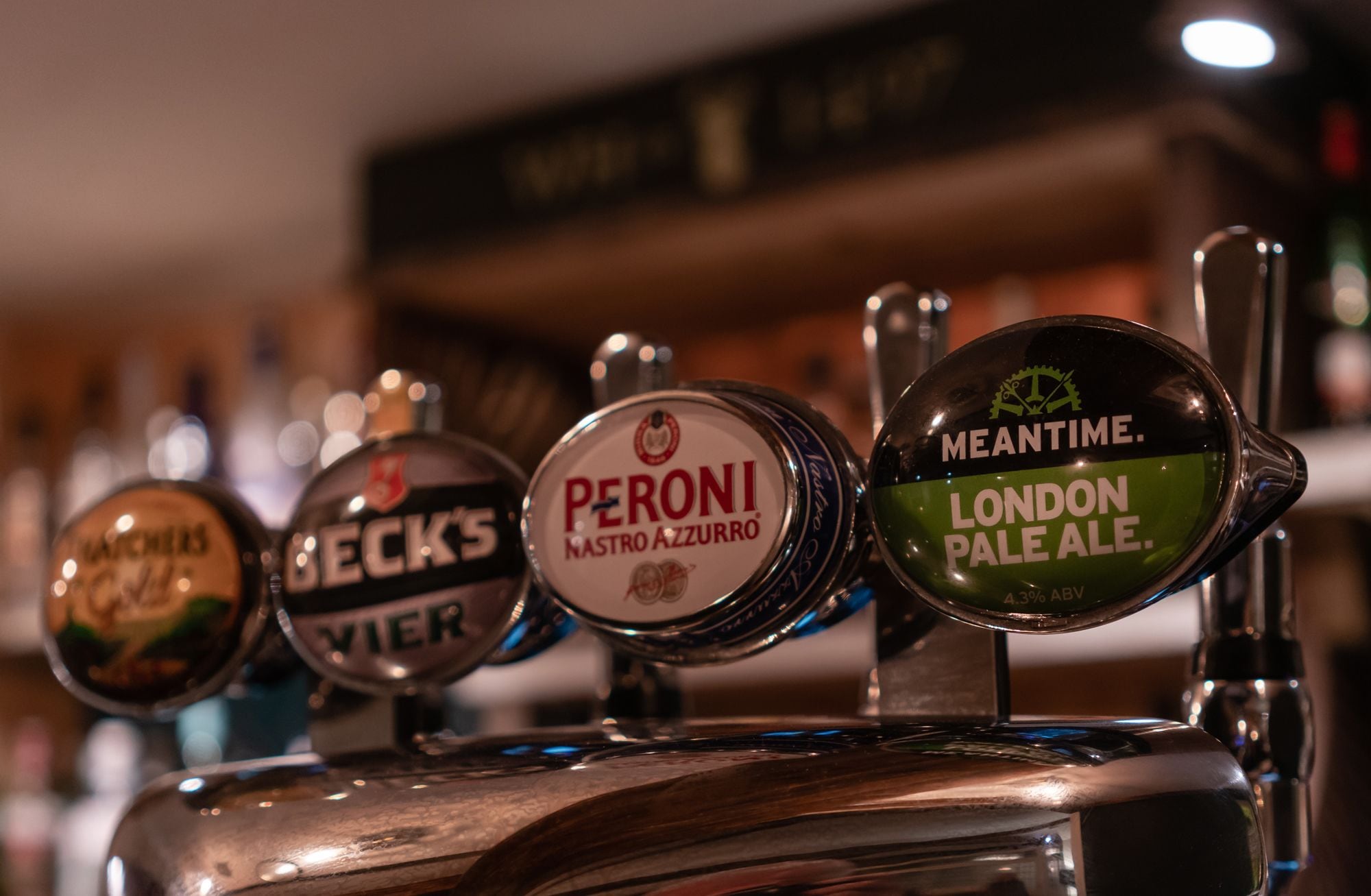 En rad med kranar för olika ölsorter, med etiketter som bland annat visar "Peroni Nastro Azzurro" och "London Pale Ale".