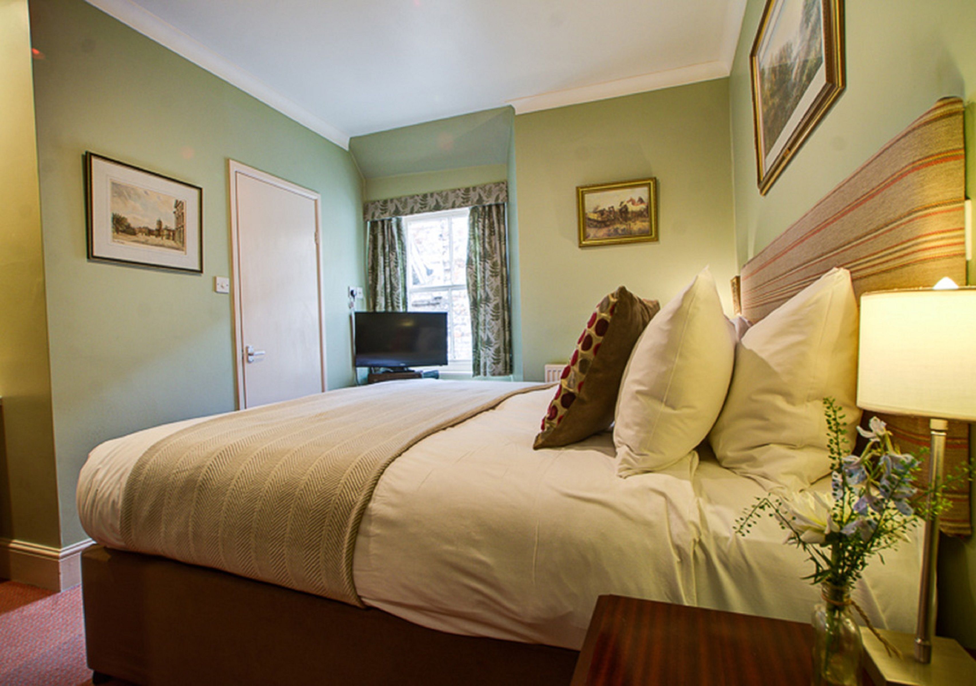 A neatly made bed with pillows, a bedside table with a lamp, and a television in the background.