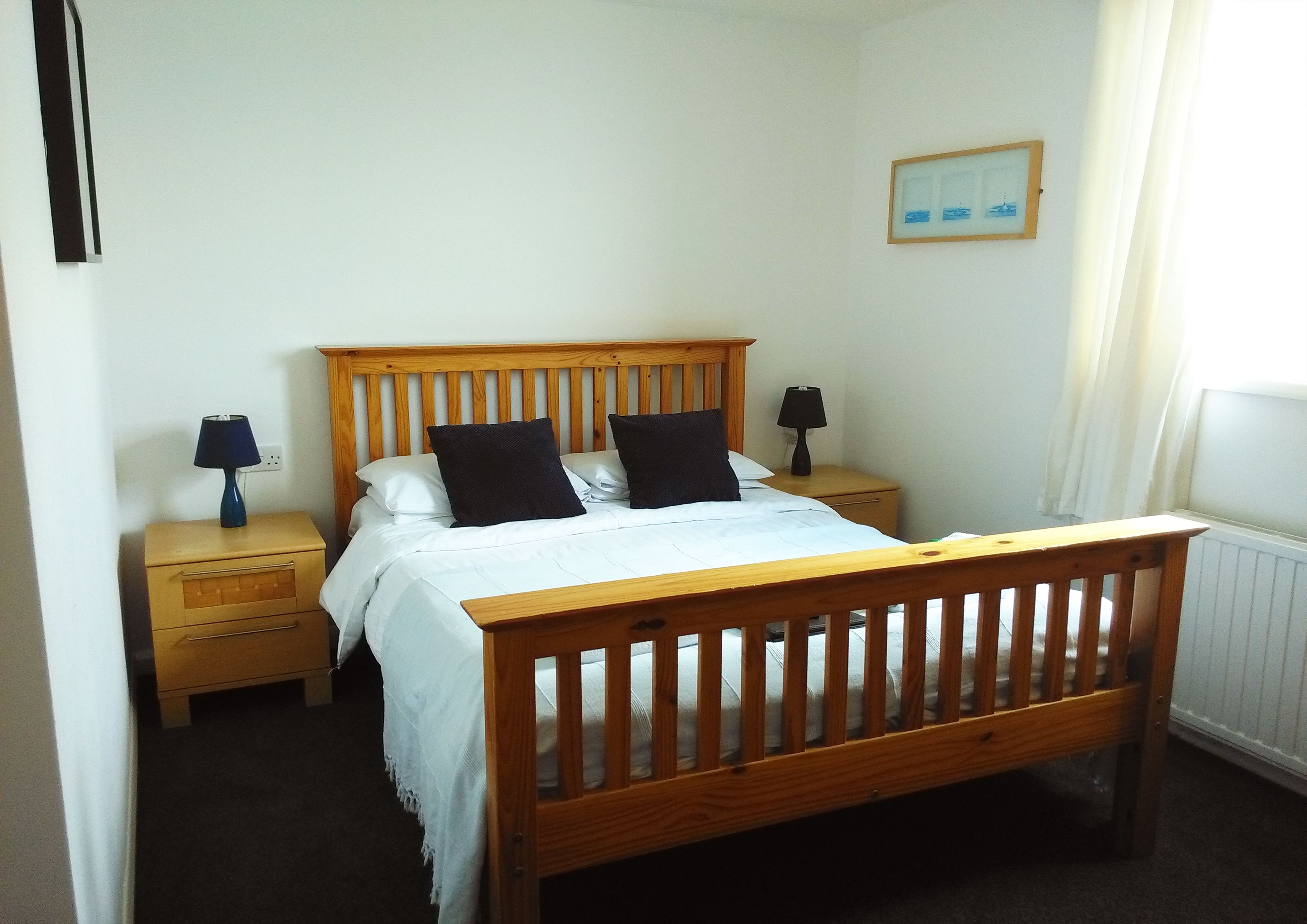Une chambre à coucher avec un lit en bois, deux tables de chevet, une commode, un tableau encadré au mur et une fenêtre avec des rideaux.