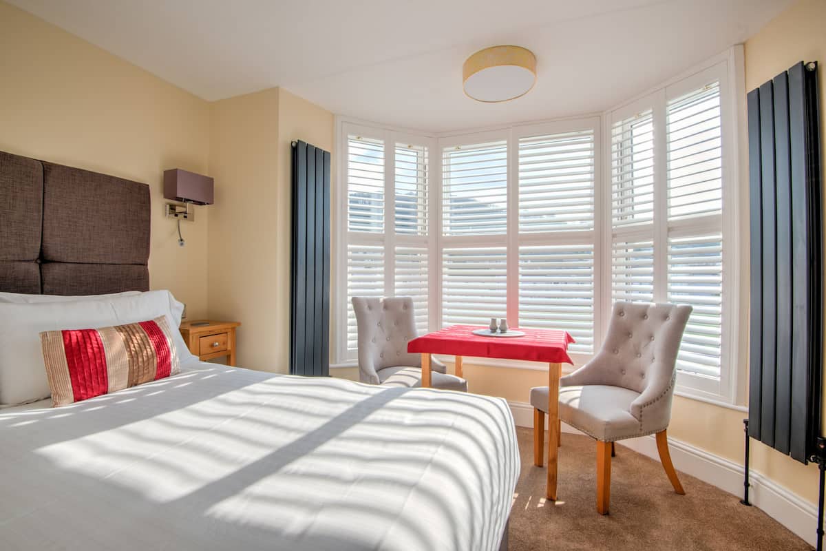 Une chambre d’hôtel avec un grand lit, une petite table, deux chaises, une lampe et une grande fenêtre avec des stores.