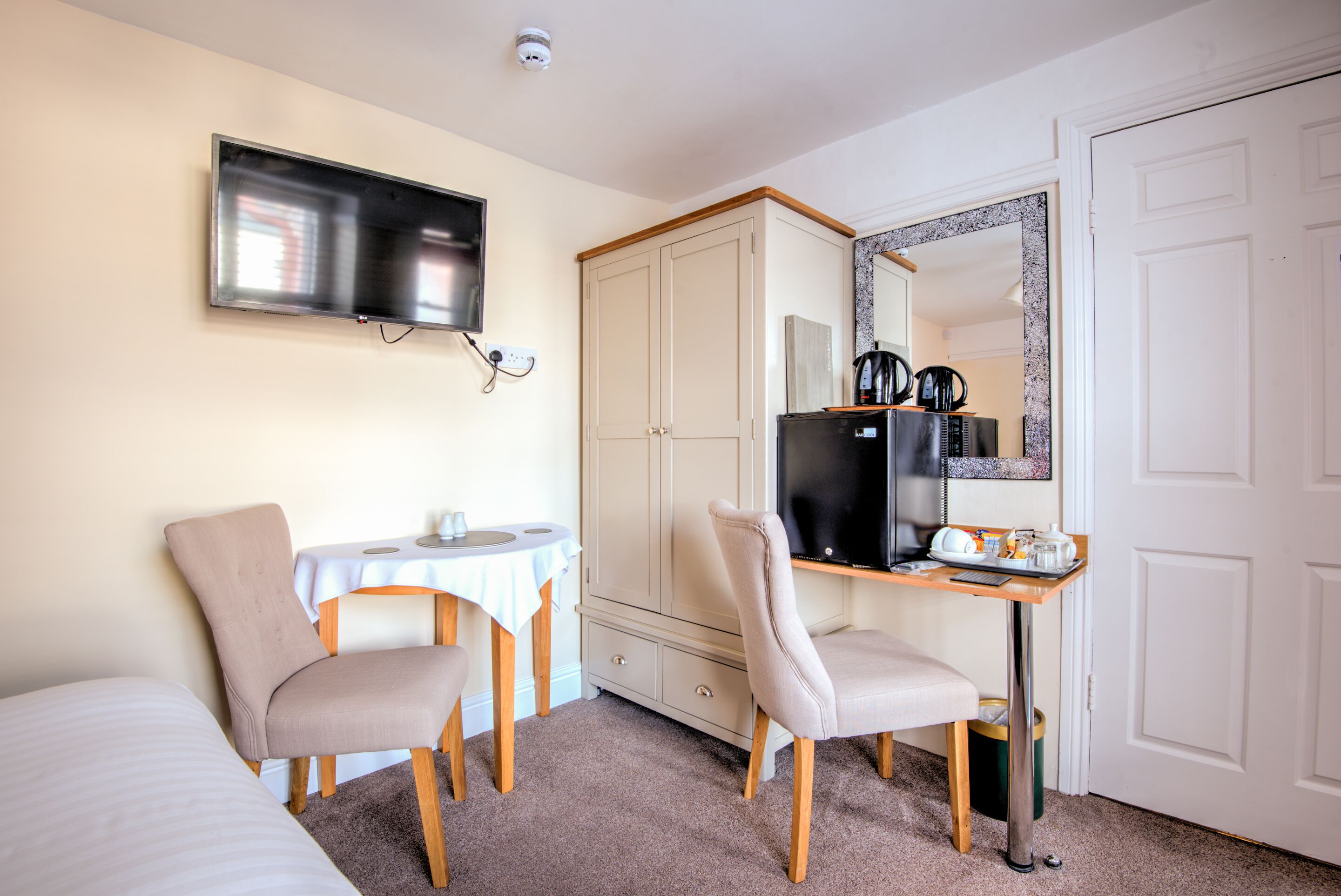A hotel room with a bed, two chairs, a desk with a computer, a television, a wardrobe, and a mirror.