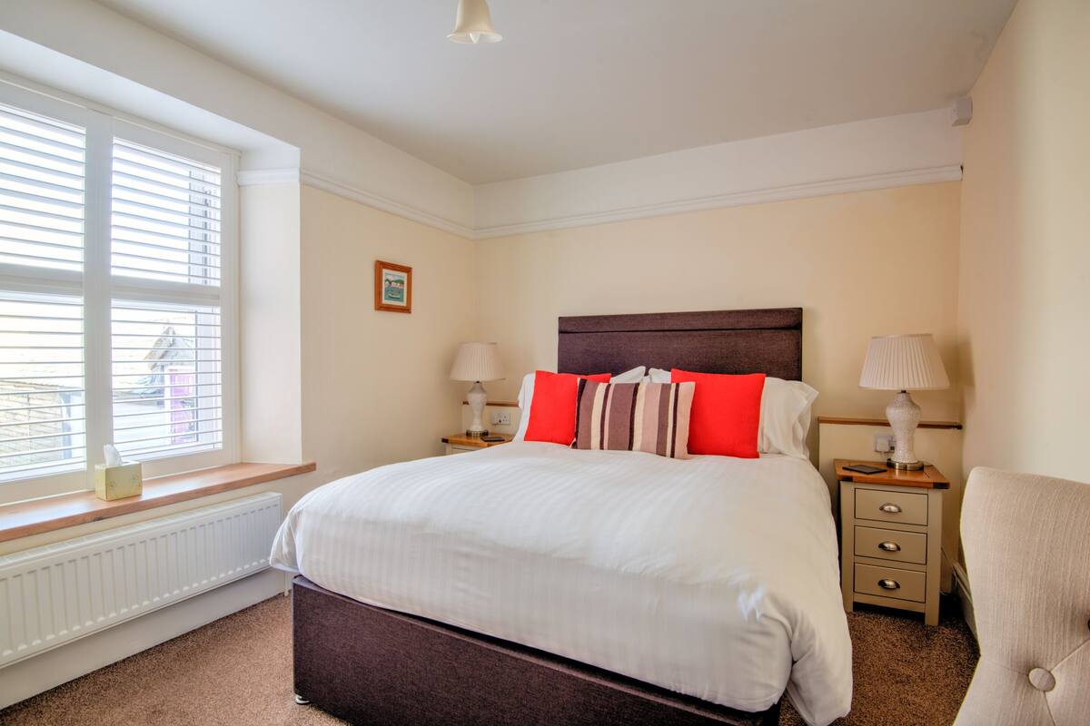 Une chambre avec un grand lit, deux tables de chevet, une chaise, une fenêtre avec des stores et un radiateur.