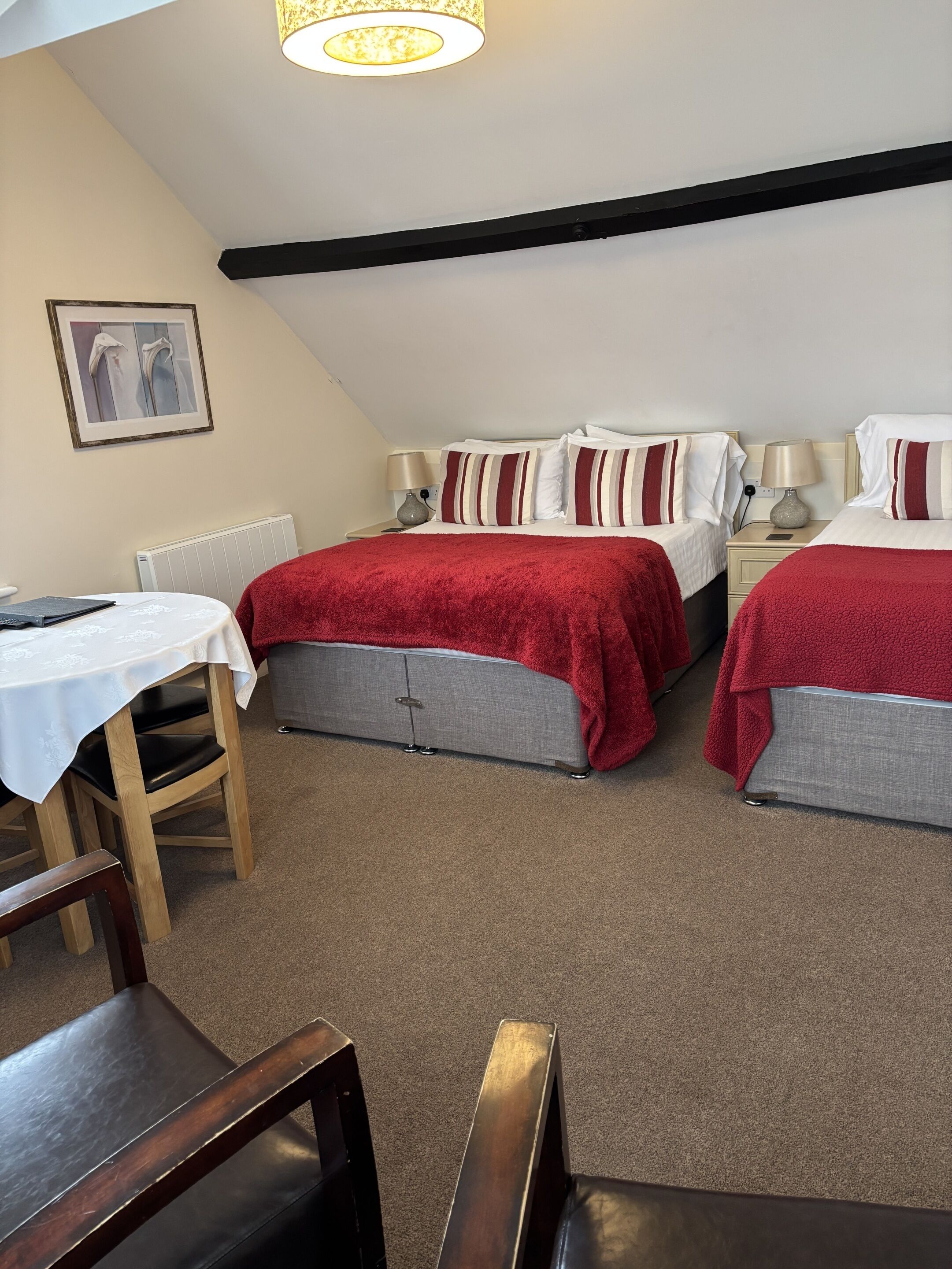 A room with two beds, a table, and chairs. There is a radiator under the table and a lamp on each bedside table.
