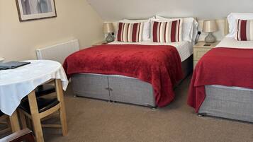 A room with two beds, a table, and chairs. There is a radiator under the table and a lamp on each bedside table.