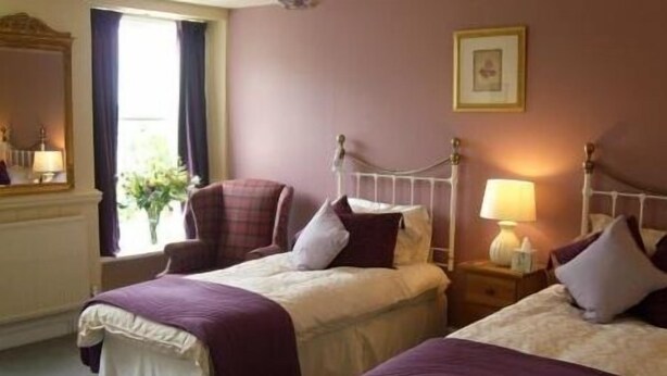 A hotel room with two beds, a chair, a nightstand with a lamp, and a window with curtains. - The Pack Horse (South Brent)