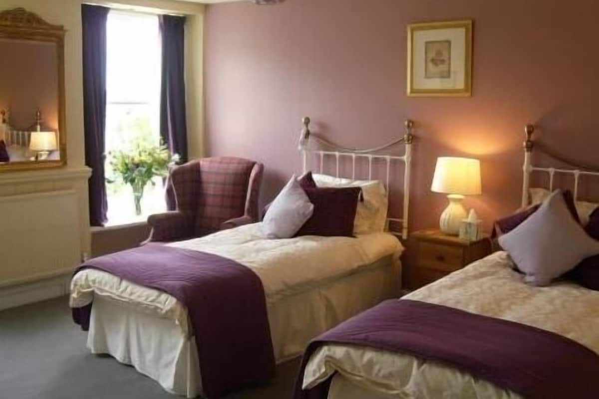 A hotel room with two beds, a chair, a nightstand with a lamp, and a window with curtains.
