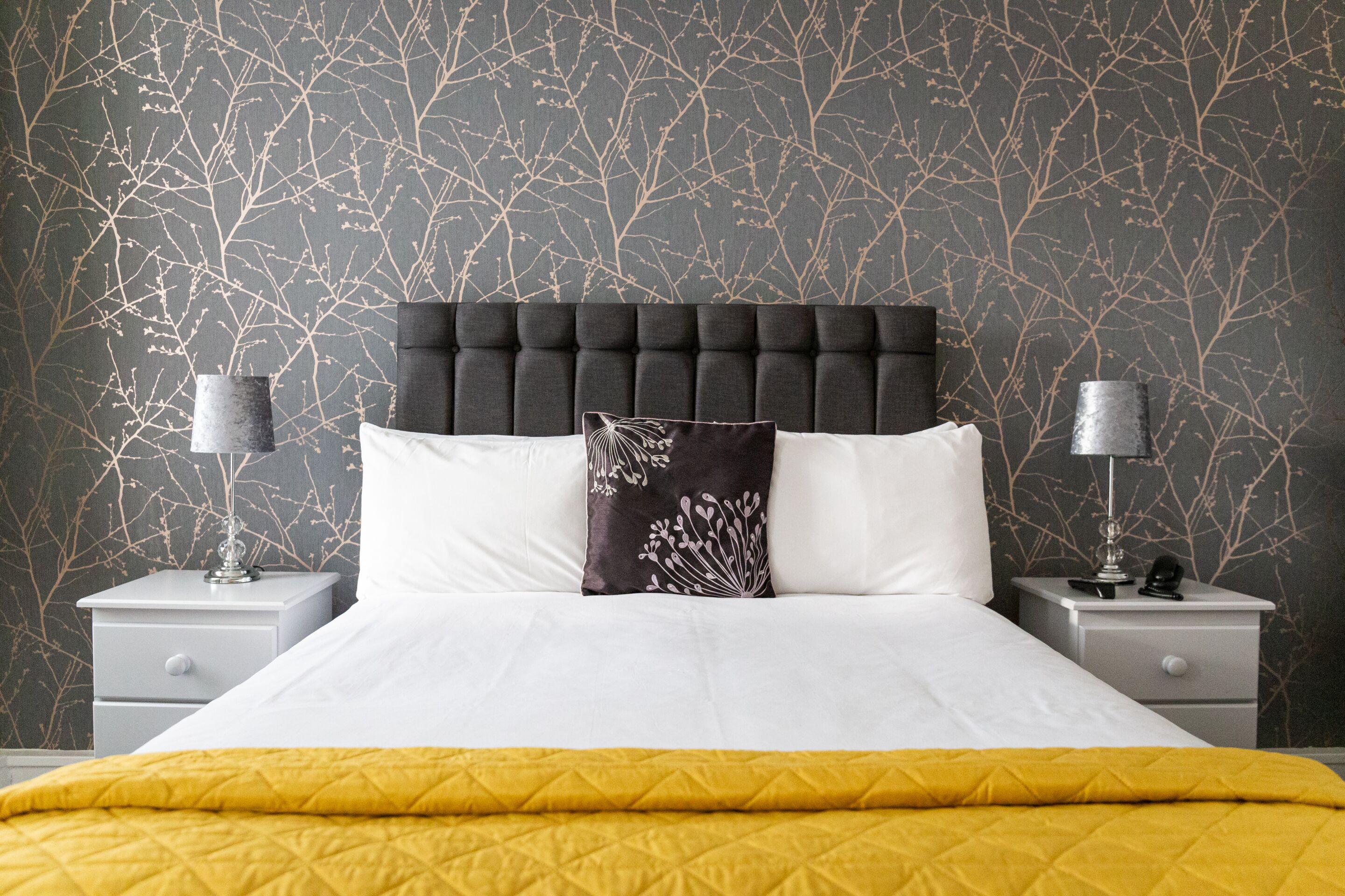 A neatly made bed with a yellow quilt, two bedside tables with lamps, and a dark headboard.
