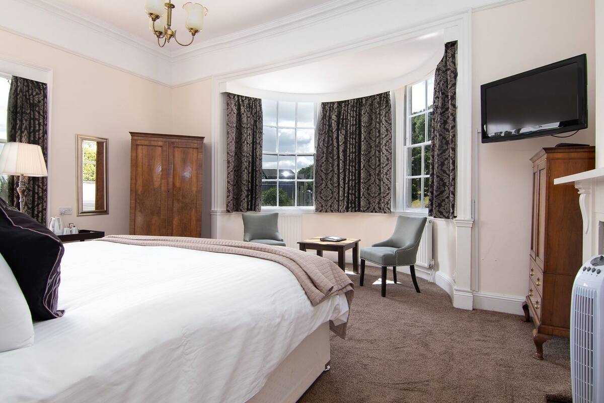 A hotel room with a large bed, a desk, two chairs, a television, and a wardrobe.