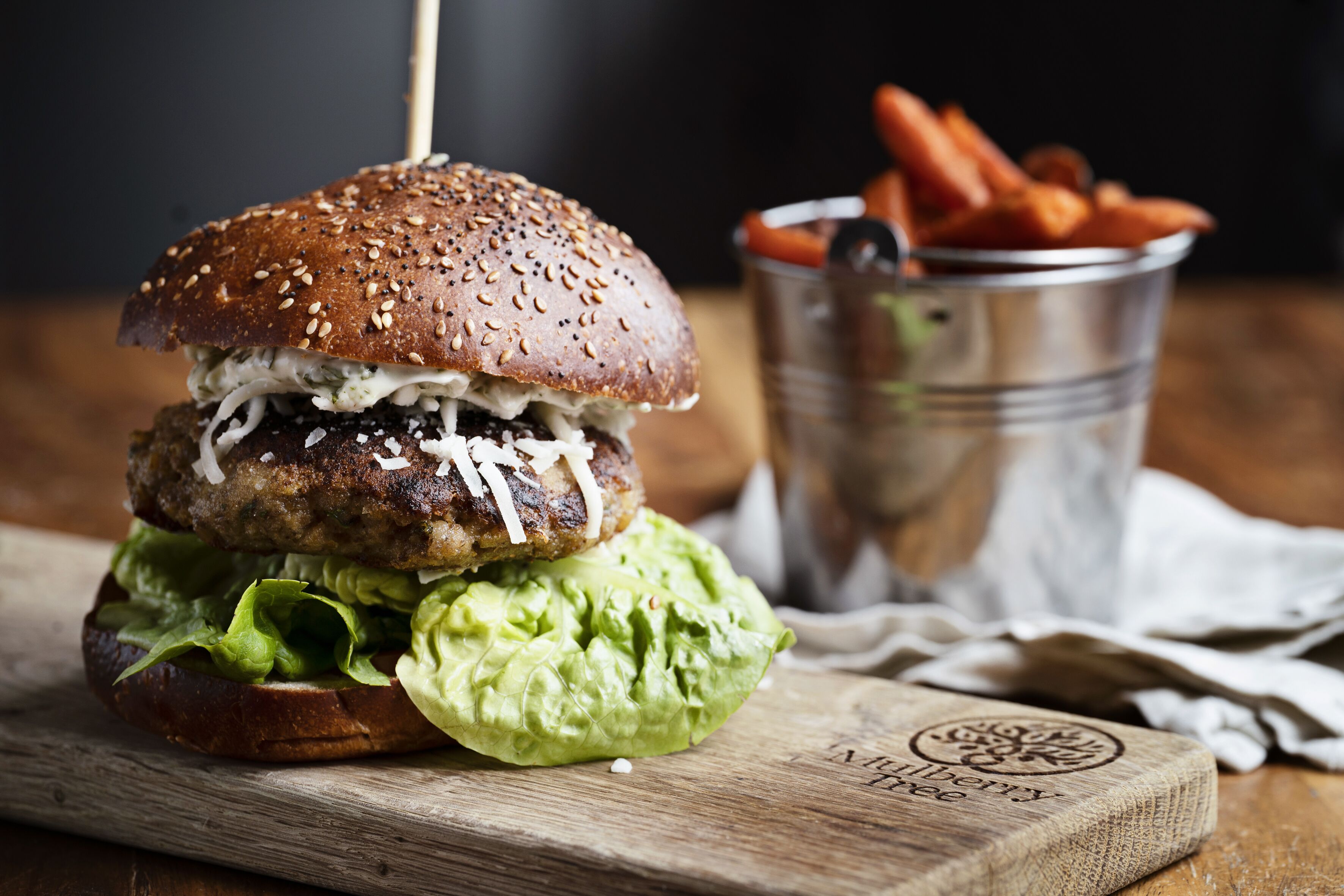 Una hamburguesa con lechuga y pan, sobre una tabla de madera, con un cubo metálico de papas fritas al fondo.