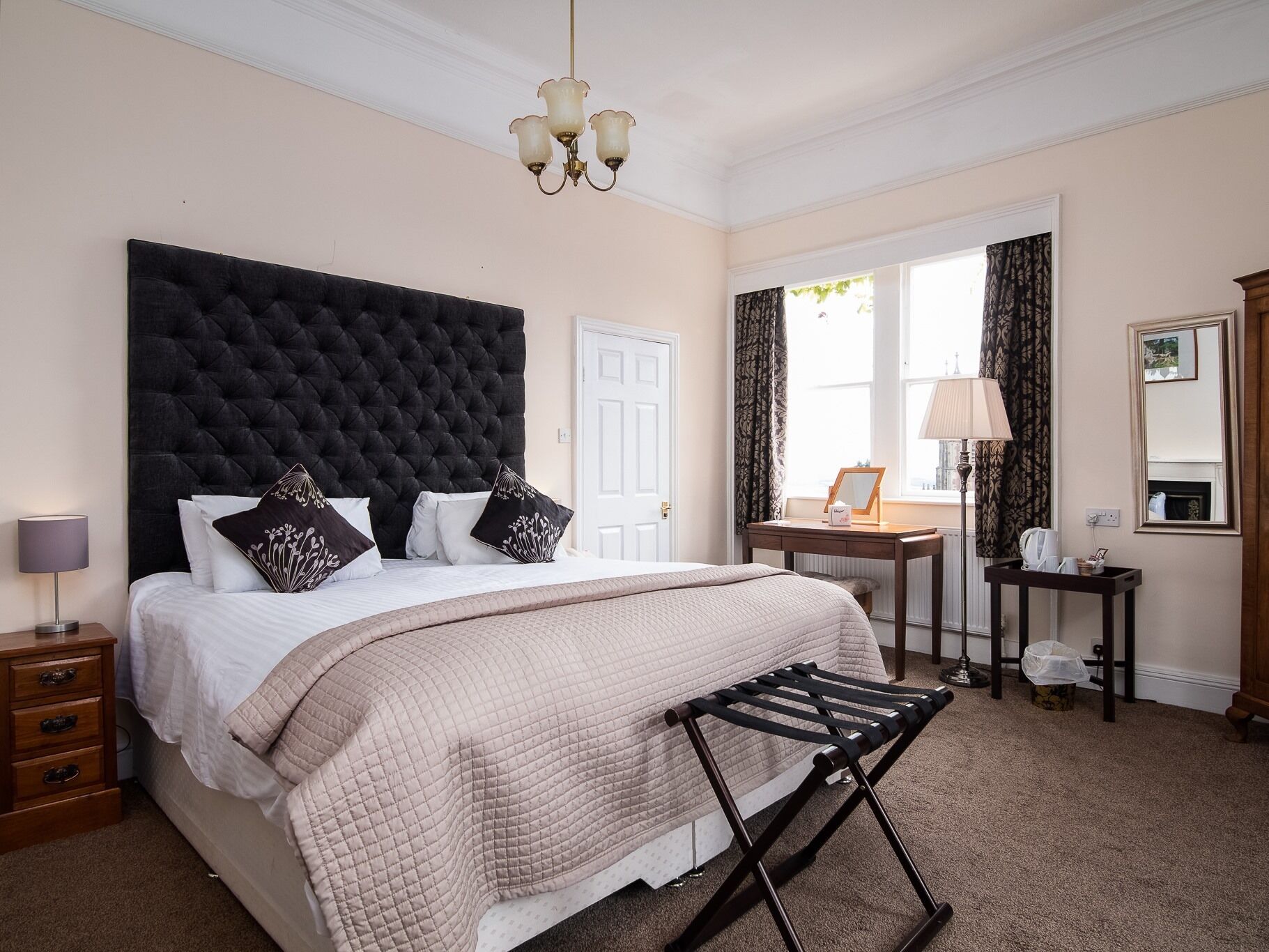 A bedroom with a large bed, a bedside table, a nightstand, a desk, a chair, a hanging lamp, a mirror, and a window with curtains.