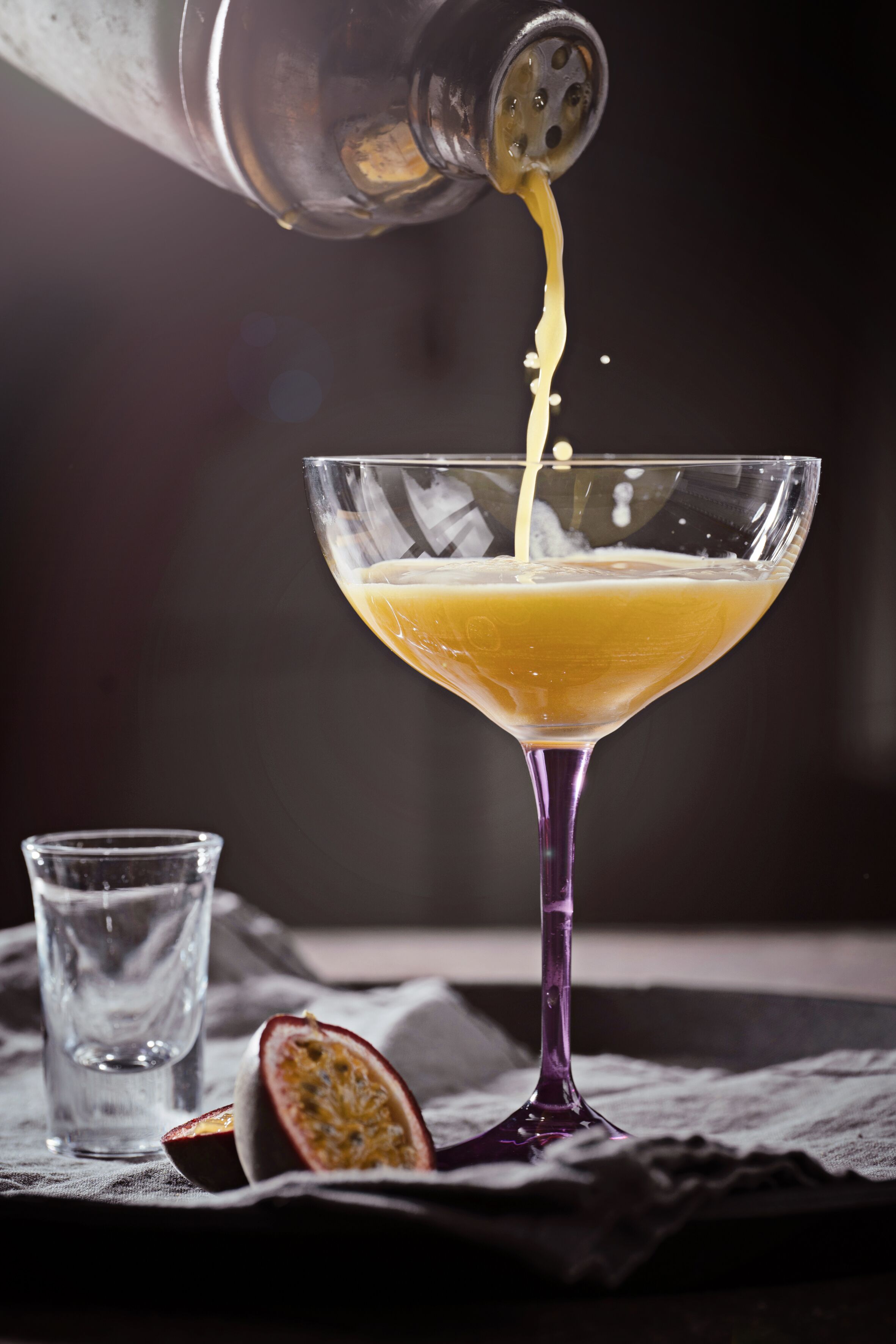 Ein Martini-Glas mit gelber Flüssigkeit, ein Cocktail-Shaker, der hineingießt, und eine halbe Passionsfrucht auf einem Tuch.