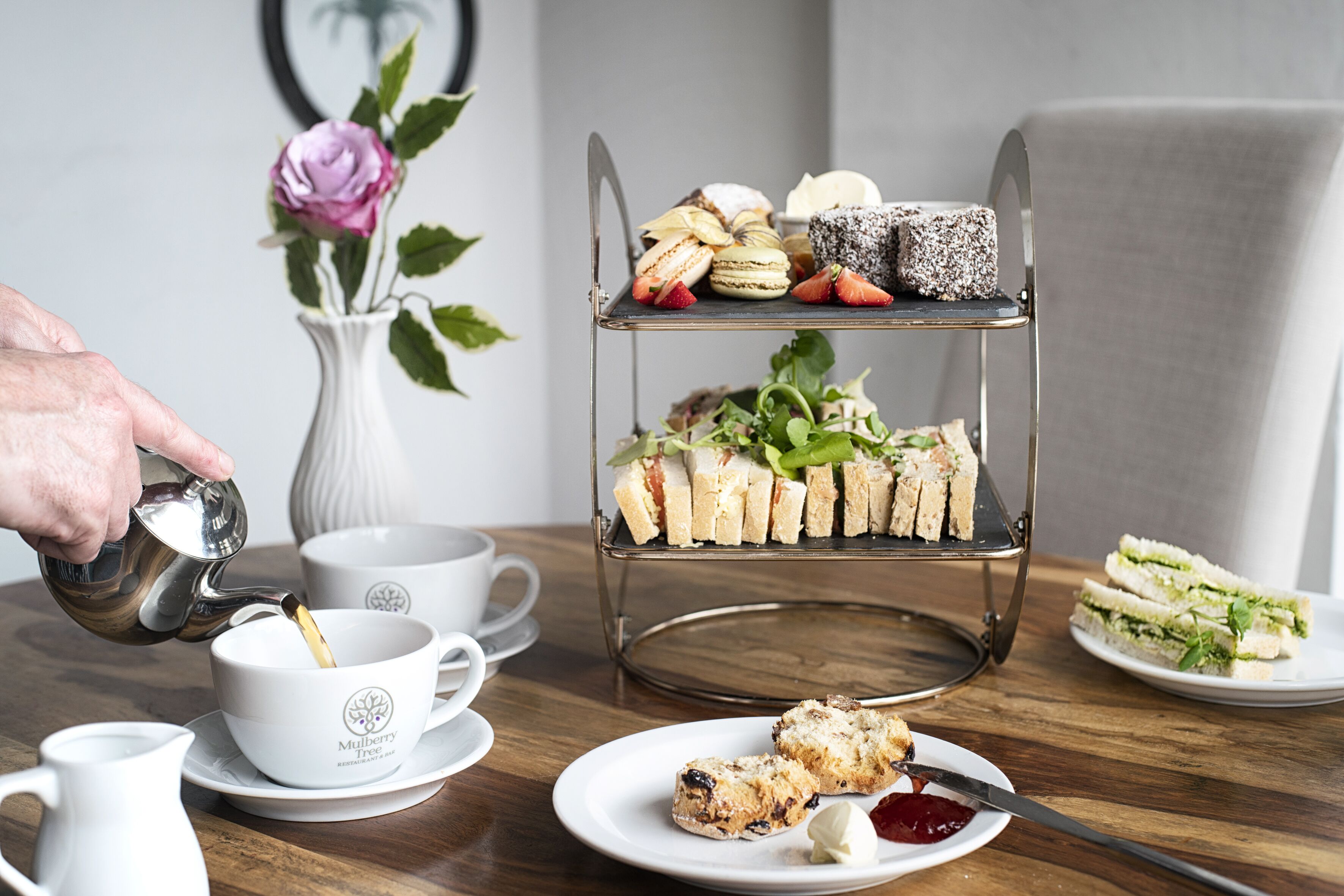 A three-tiered dessert stand with various pastries and sandwiches, a white teapot pouring tea, and a floral arrangement in the background.