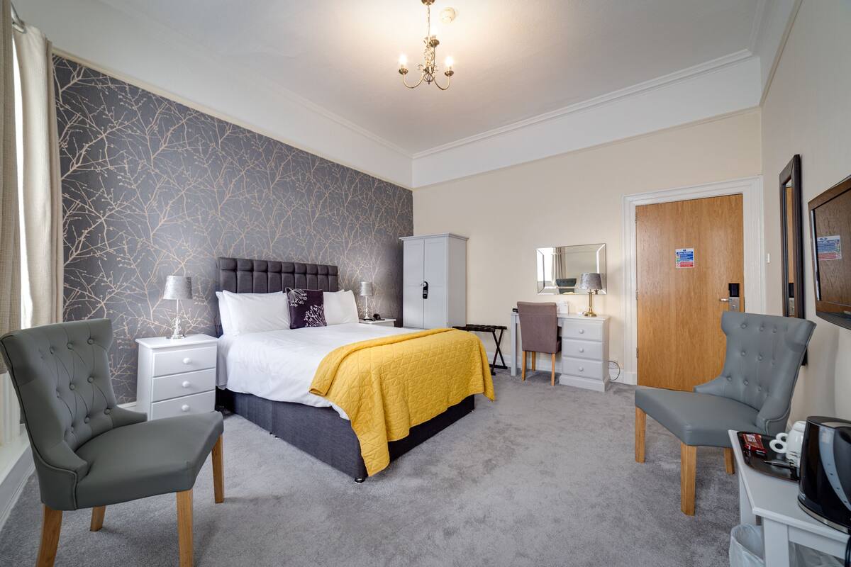 A bedroom with a bed, two armchairs, a desk, a wardrobe, and a mirror.