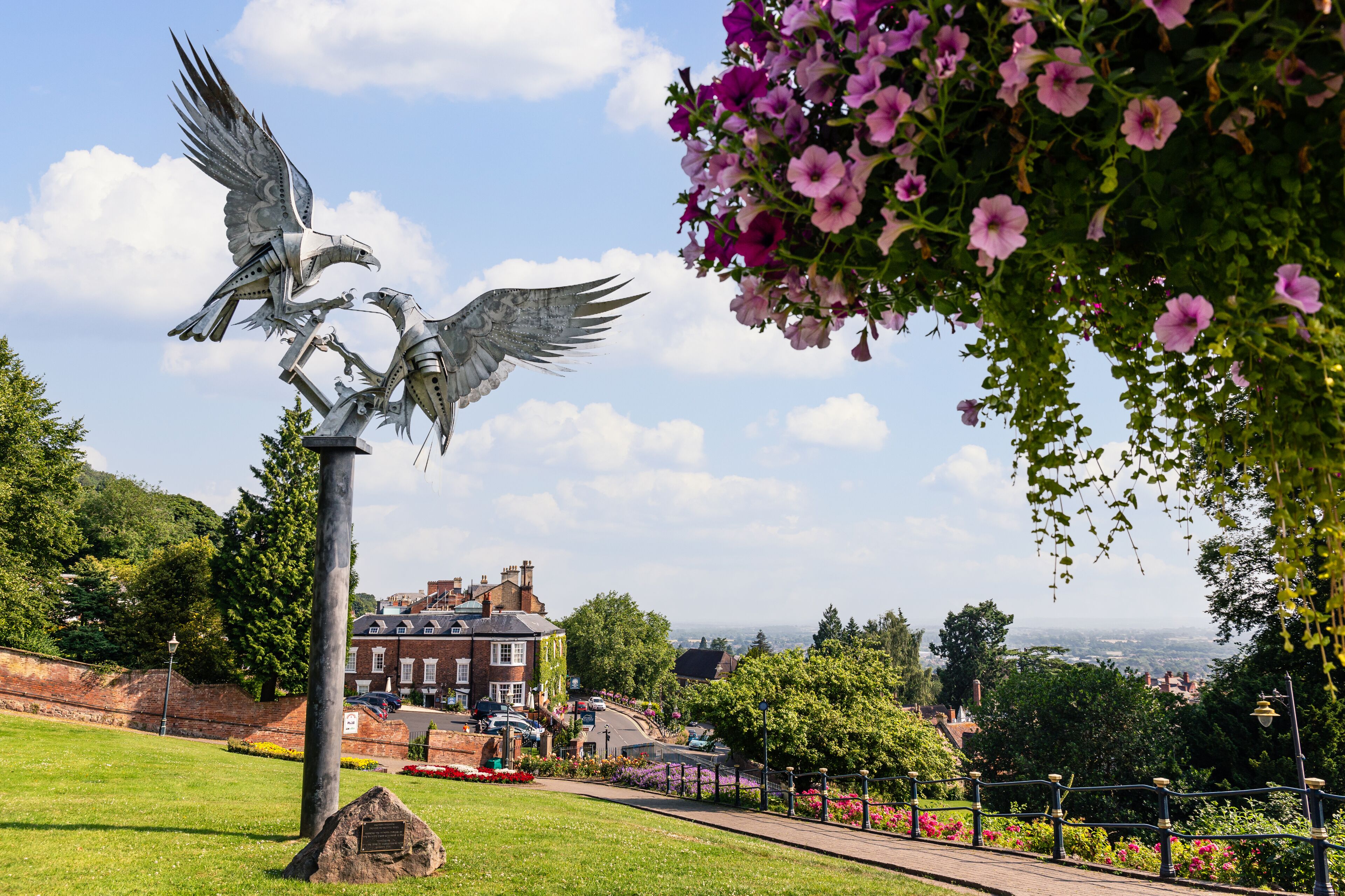 A park with a metal sculpture of birds, a brick building, and a view of the city.