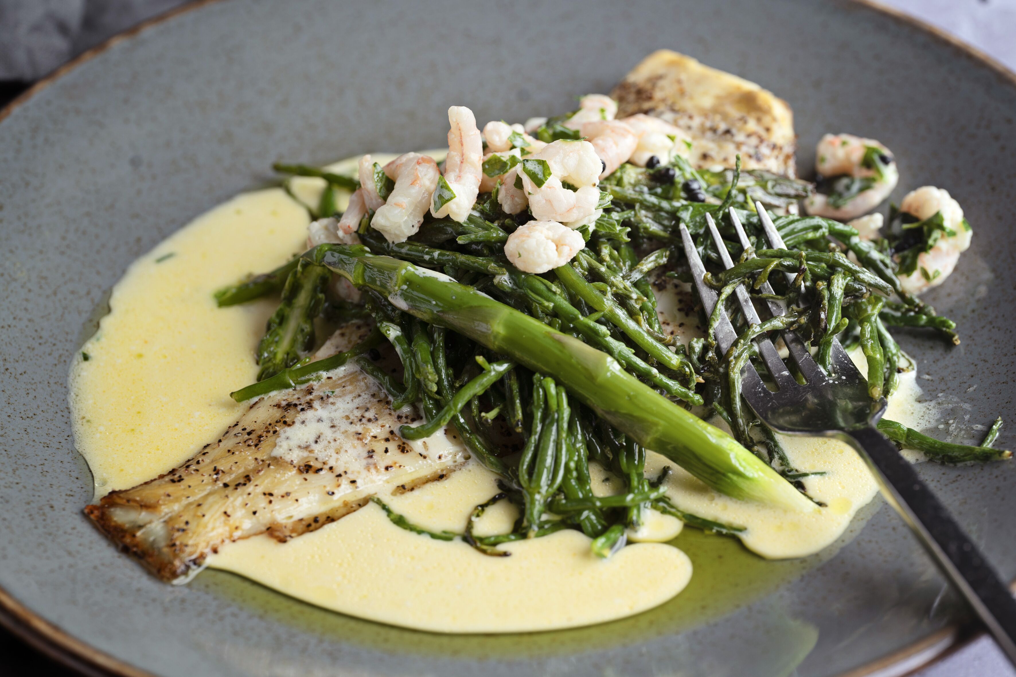 Un piatto di cibo con gamberi, asparagi e un pezzo di pesce, accompagnato da una salsa cremosa.