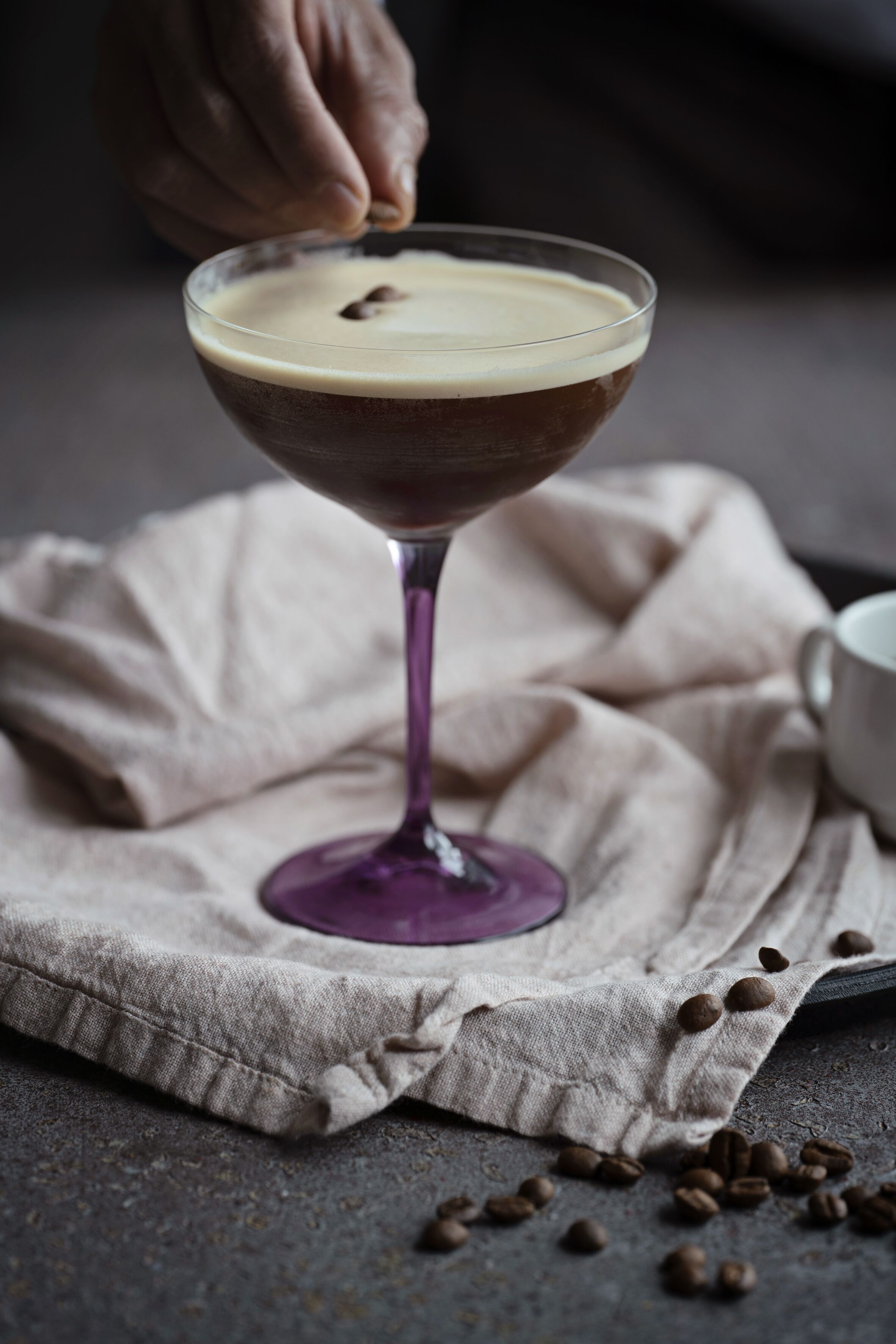 Una copa de martini con una capa cremosa, sobre un mantel con granos de café esparcidos alrededor.