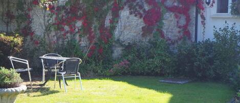 A garden with a stone wall covered in red and green foliage, a metal table and chairs, and a small window with a lantern.