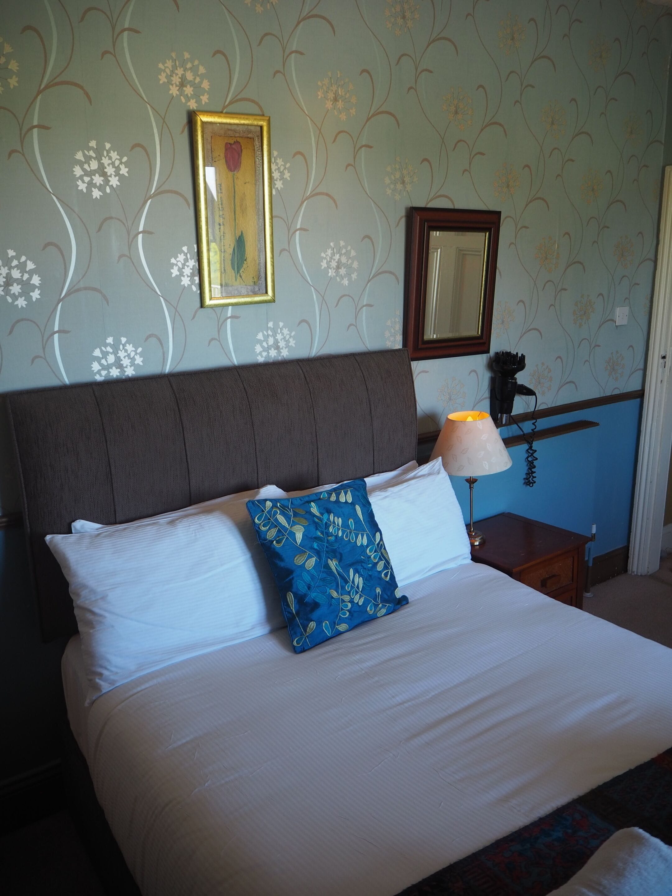 A neatly made bed with white linens and a blue floral pillow, a bedside lamp, a framed picture on the wall, and a mirror.