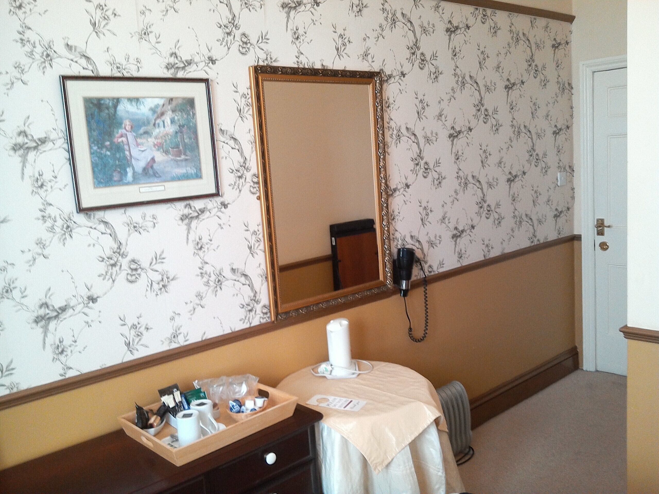 A hotel room with a floral wallpaper, a large mirror, a framed picture, a bedside table with toiletries, and a radiator.