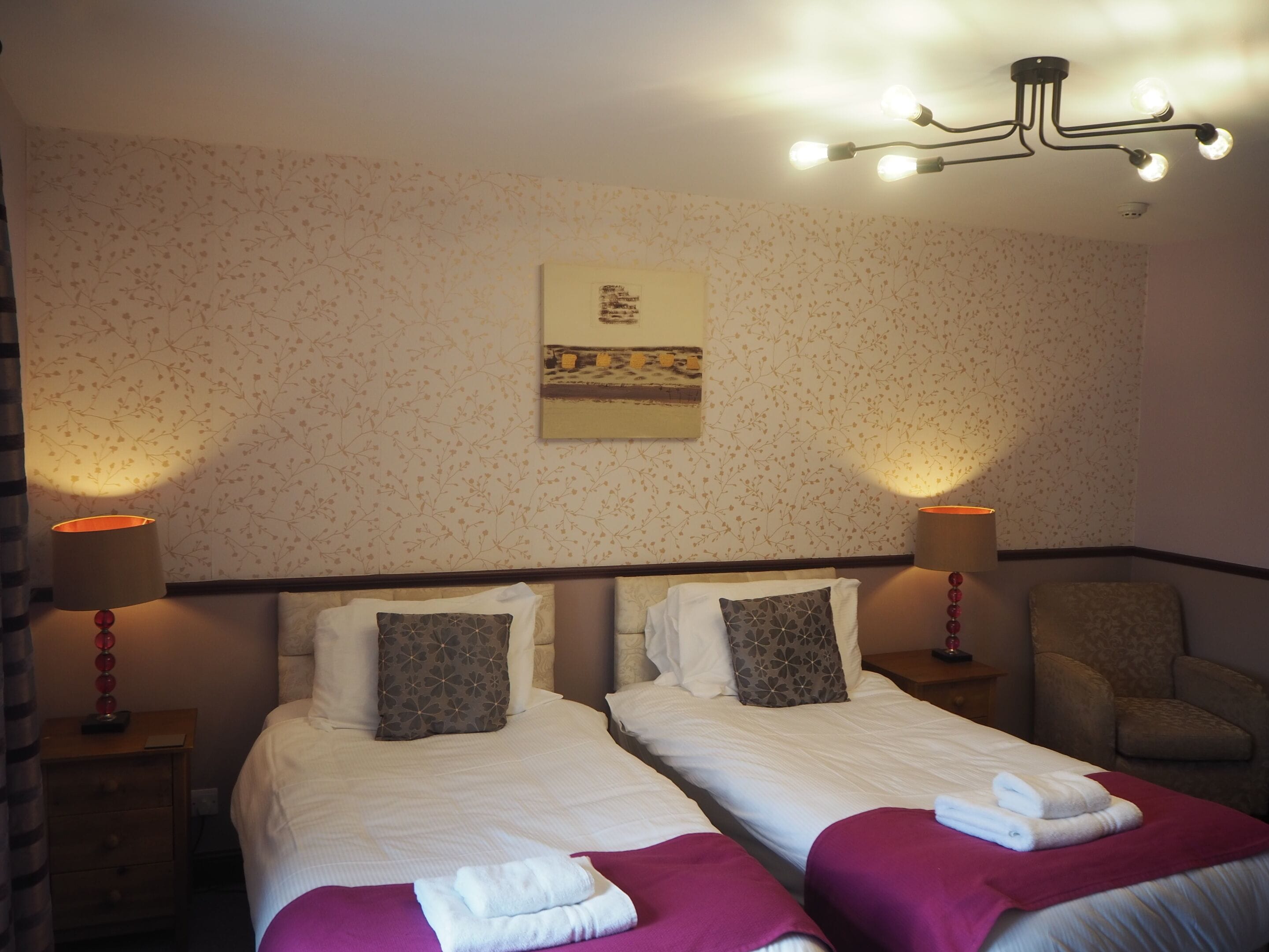 A hotel room with two beds, a chair, a lamp, and a painting on the wall.
