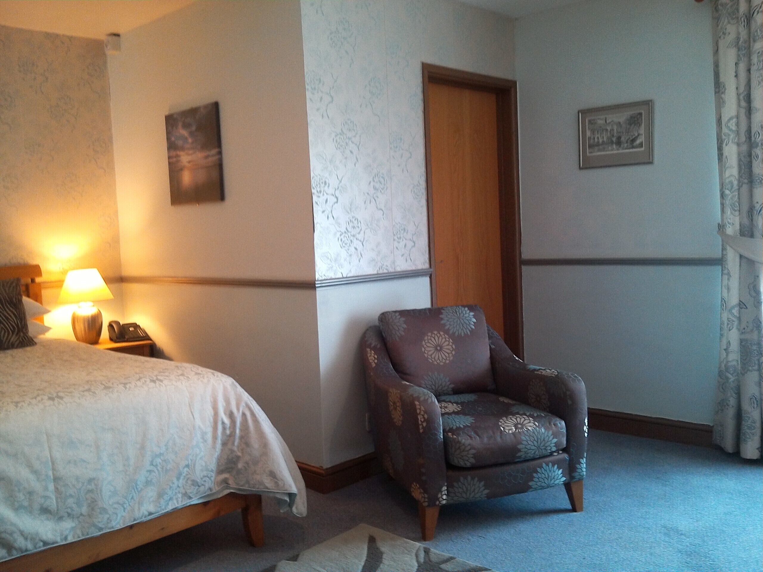 A hotel room with a bed, a chair, a nightstand with a lamp, and a door.