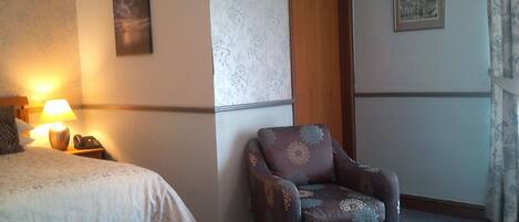 A hotel room with a bed, a chair, a nightstand with a lamp, and a door.