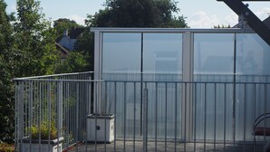 En glasindrammet balkon med metalgelændere, plantekasser og udsigt til træer og bygninger i baggrunden.
