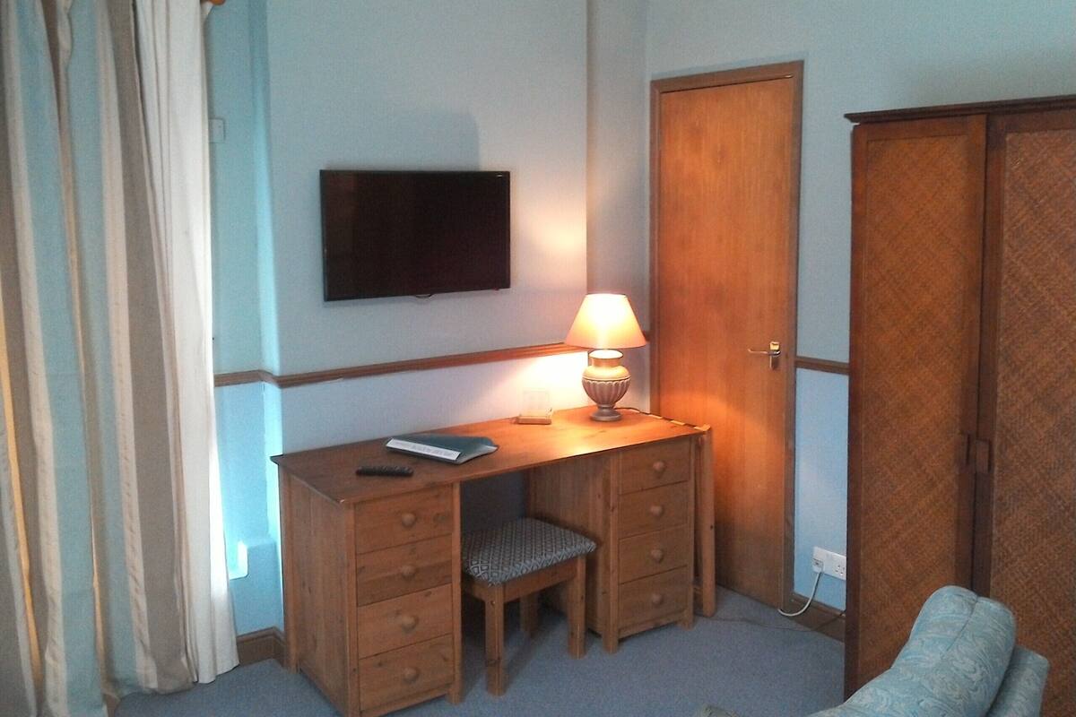 Una habitación pequeña y sencilla con un escritorio de madera, una silla, un televisor montado en la pared y un armario.