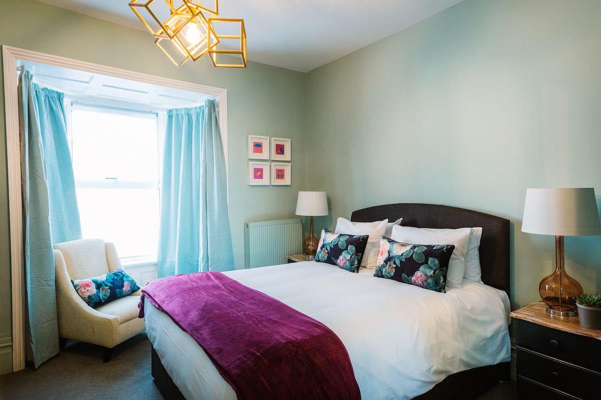 A bedroom with a large bed, a chair, a nightstand, a lamp, and a window with curtains.