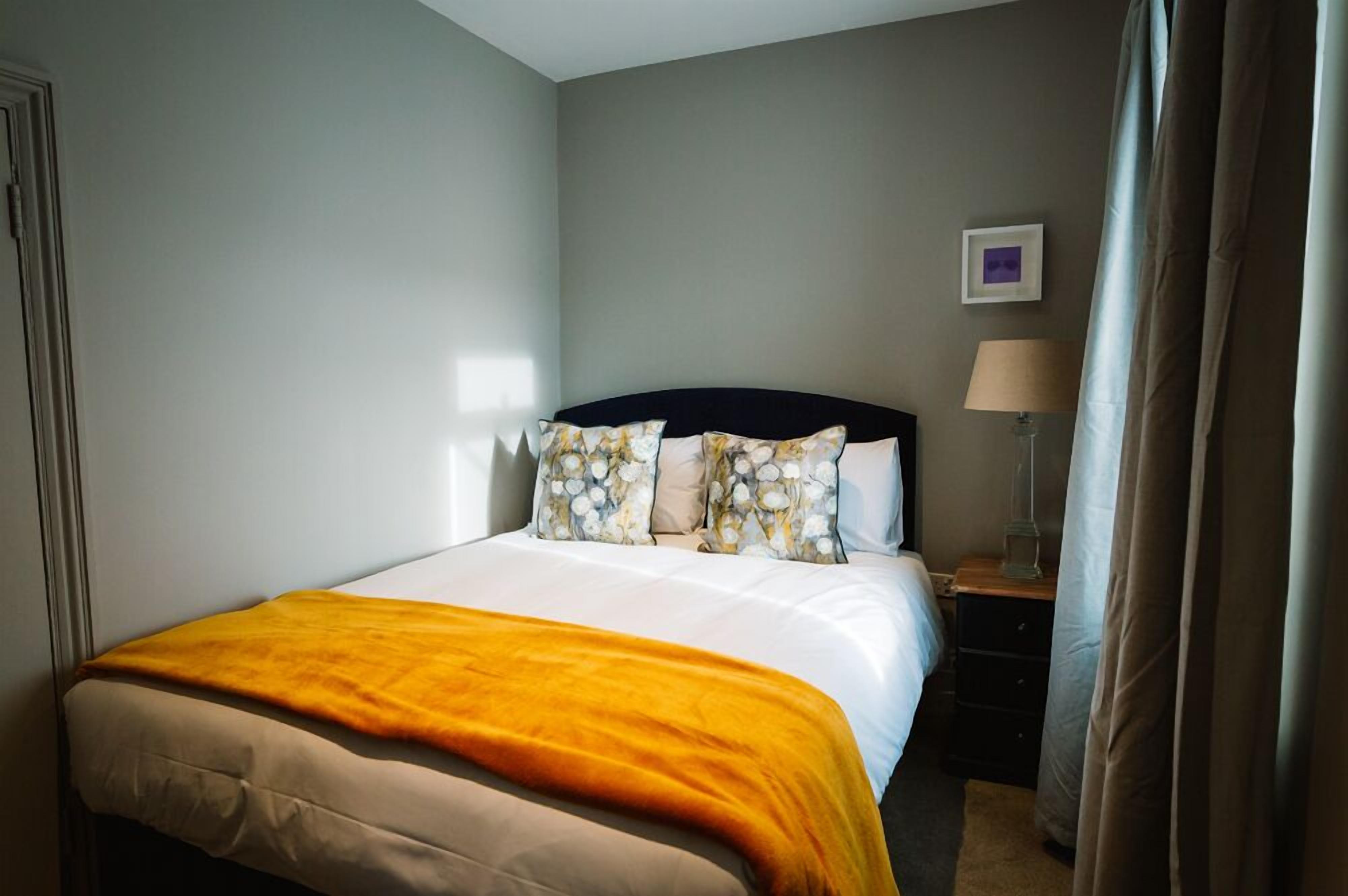 A neatly made bed with a yellow throw blanket, a bedside lamp, and a framed picture on the wall.