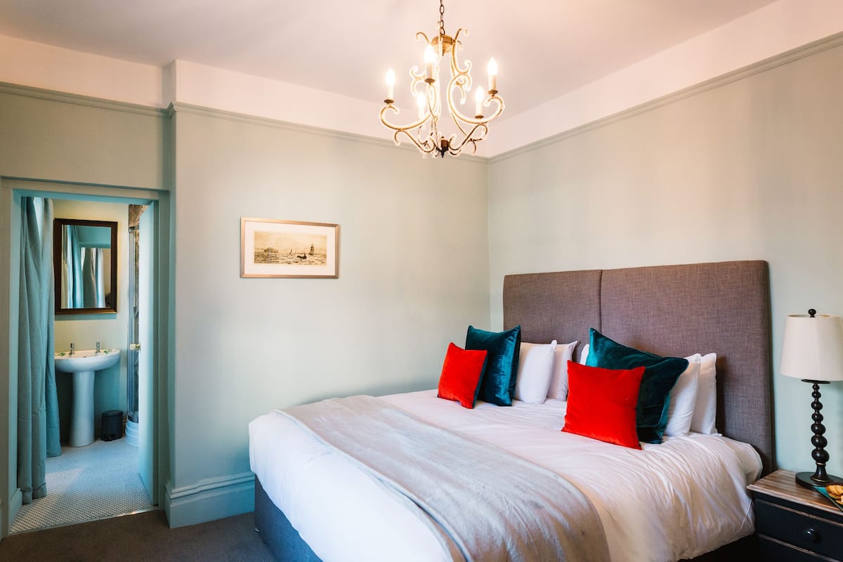 A bedroom with a large bed, a bedside table with a lamp, a framed picture on the wall, and a bathroom visible through an open door.