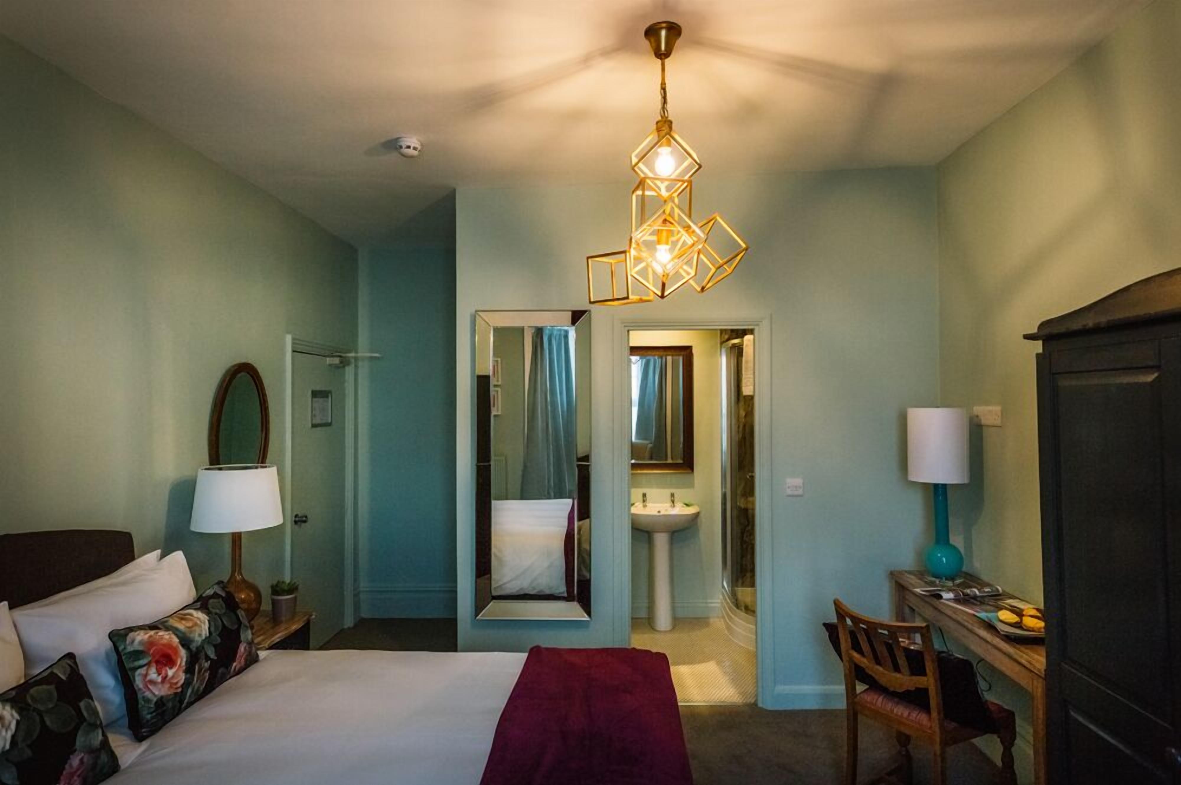 A hotel room with a bed, bedside table, lamp, mirror, and a desk with a chair.