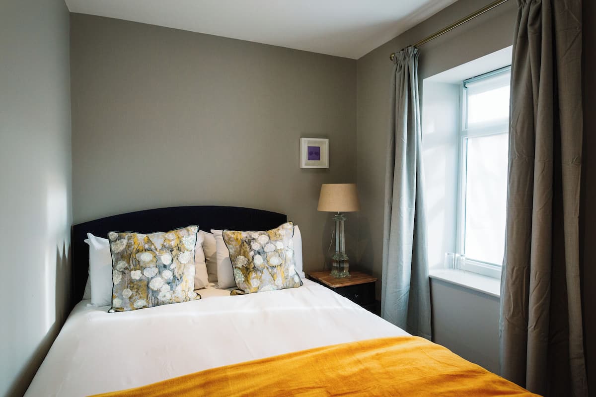 A neatly made bed with a white duvet and a yellow throw, a bedside table with a lamp, and a window with curtains.