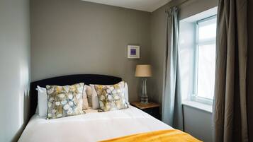 Una cama bien tendida con edredón blanco y manta amarilla, una mesita de noche con lámpara, y una ventana con cortinas.