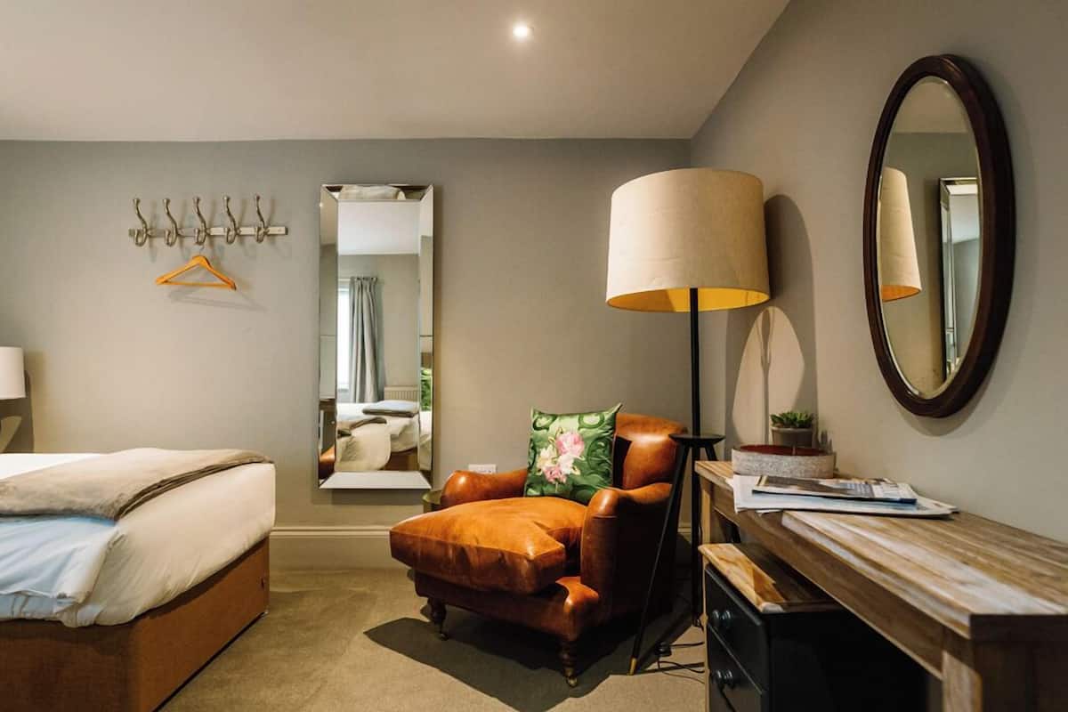 A bedroom with a bed, a chair, a desk, a lamp, a mirror, and a coat rack.